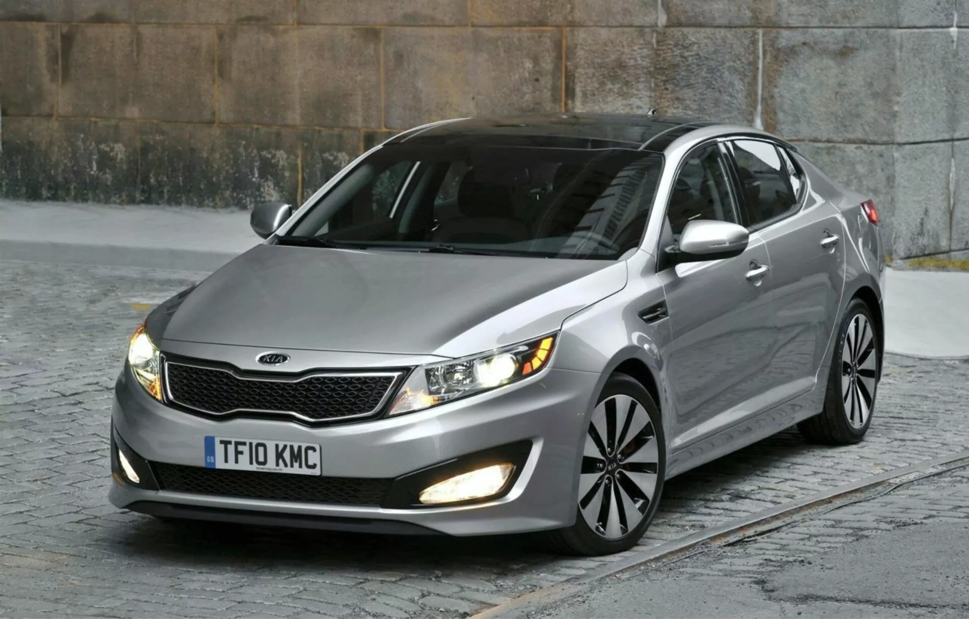 Kia Optima Limousine 2012 garée sur des pavés capturant son quart avant gauche y compris la grille avec accents chromés, phares à lentille claire, feux de brouillard, peinture métallique, roues en alliage à rayons multiples, et miroirs avec indicateurs de signal intégrés, soulignant son design aérodynamique et les caractéristiques de berlines modernes dans un cadre urbain