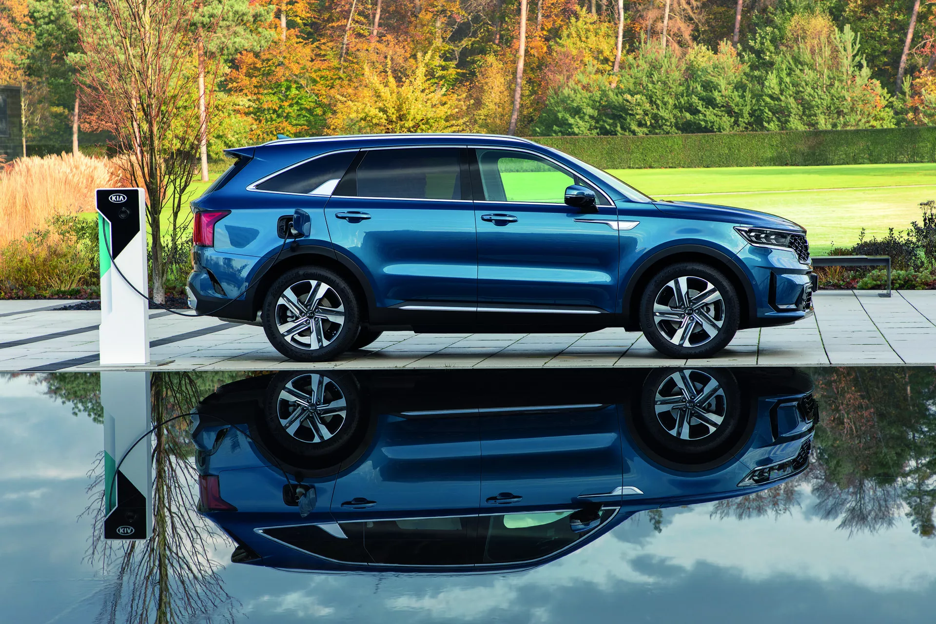 SUV bleu moderne Kia Sorento MQ4 2020 garé sur un pavé gris à côté d'une station de recharge pour véhicule électrique, montrant le profil latéral avec une vue partielle de l'avant, mis en évidence par une lumière naturelle du matin ou du soir avec un fond de feuillage vert, soulignant la capacité de recharge électrique, le design aérodynamique et les roues en alliage.