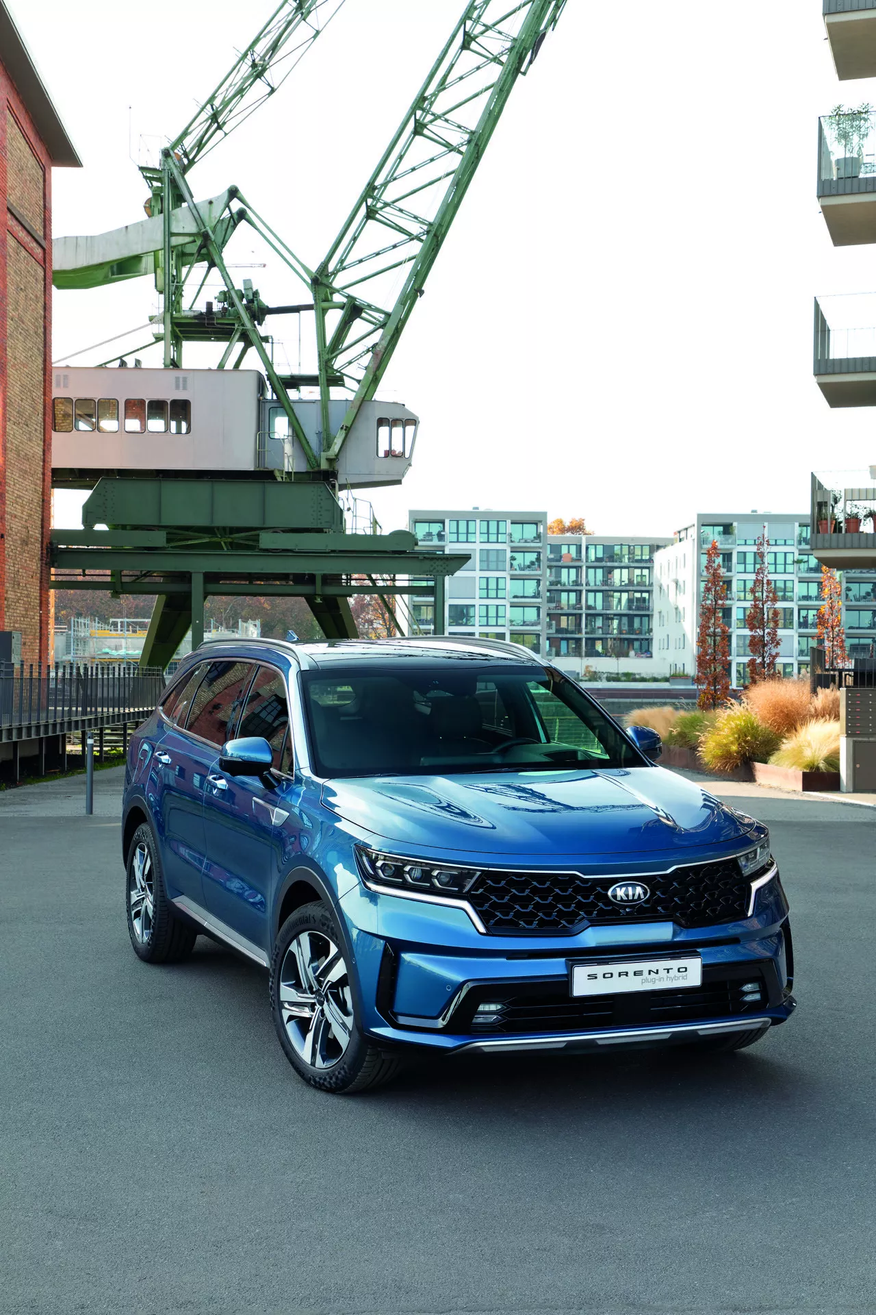 Kia Sorento MQ4 2020 en bleu présentée dans un environnement urbain avec une grue industrielle et des bâtiments modernes en arrière-plan, mettant en lumière les phares LED, la grille géométrique avec de potentiels capteurs ADAS, les roues en alliage à multiples branches et les clignotants intégrés dans les rétroviseurs latéraux, soulignant ses caractéristiques technologiques et son apparence sportive.