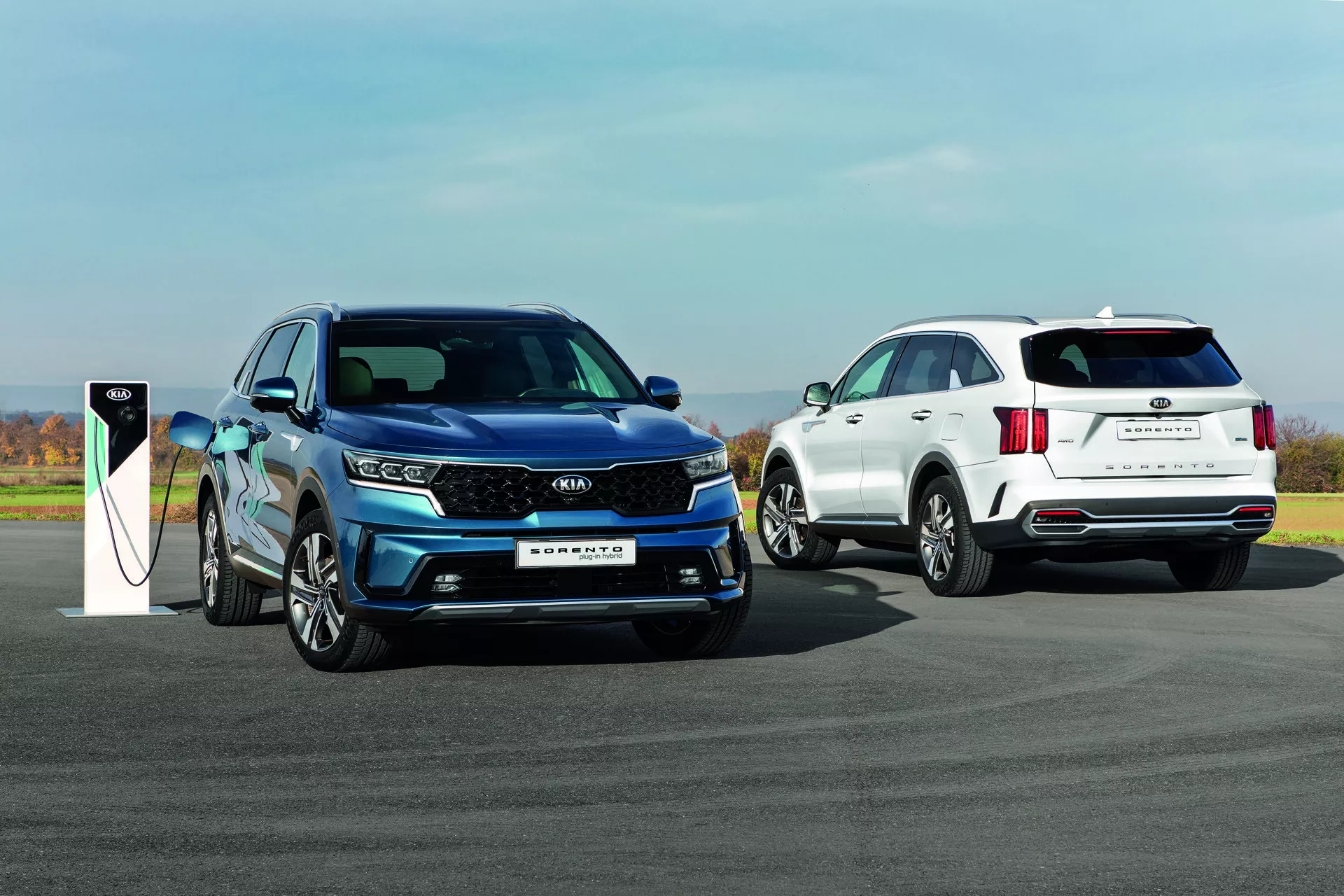 Kia Sorento MQ4 2020 en bleu, modèle électrique ou hybride rechargeable en charge, et un SUV blanc en arrière-plan sur une surface extérieure pavée sous un ciel dégagé