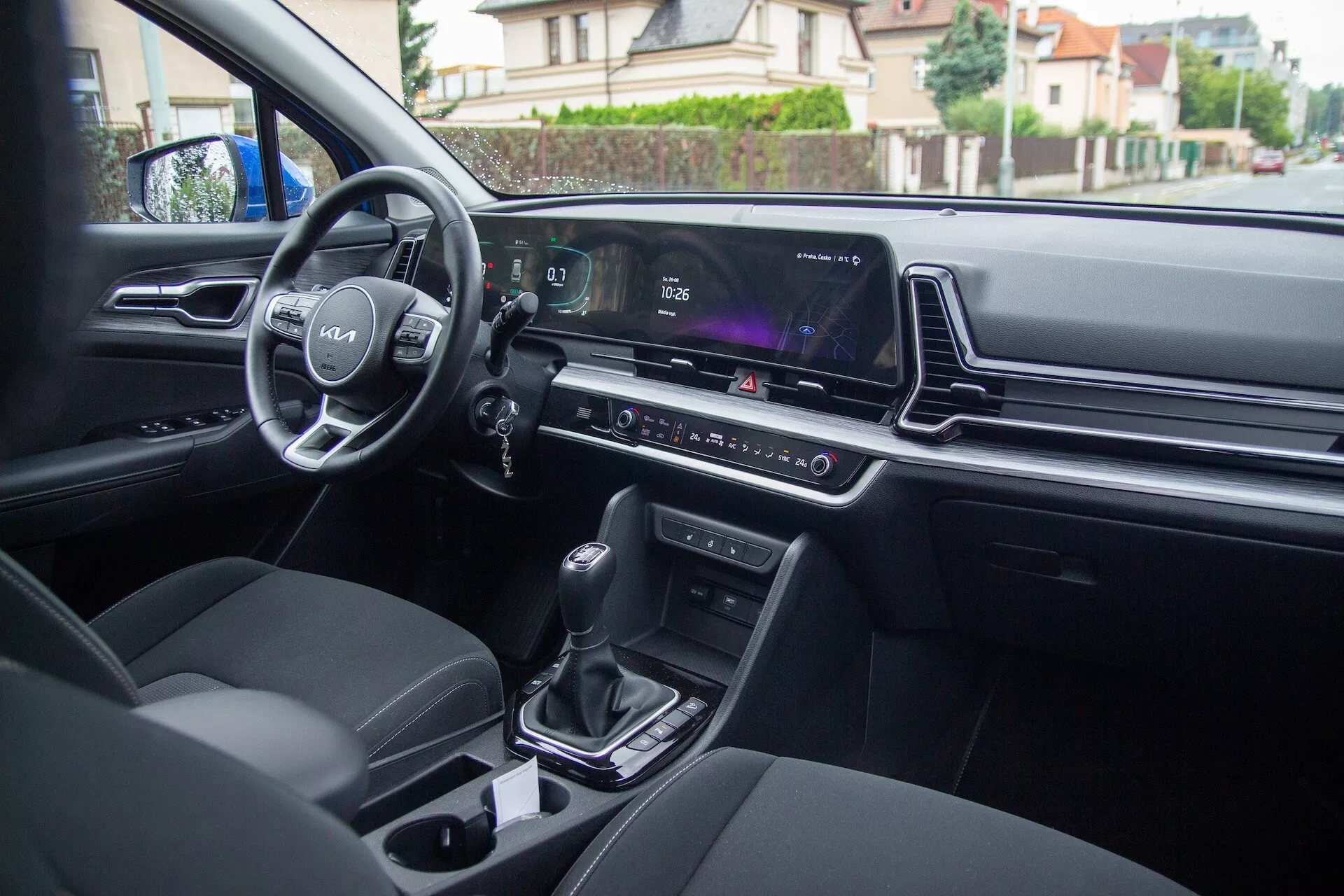 Vue intérieure de la Kia Sportage 2022 depuis le siège arrière montrant les commandes sur la porte du conducteur, le volant avec logo et commandes montées, le tableau de bord numérique, l'écran d'infodivertissement, le contrôle climatique, le levier de vitesse automatique, les porte-gobelets et les sièges modernes rembourrés