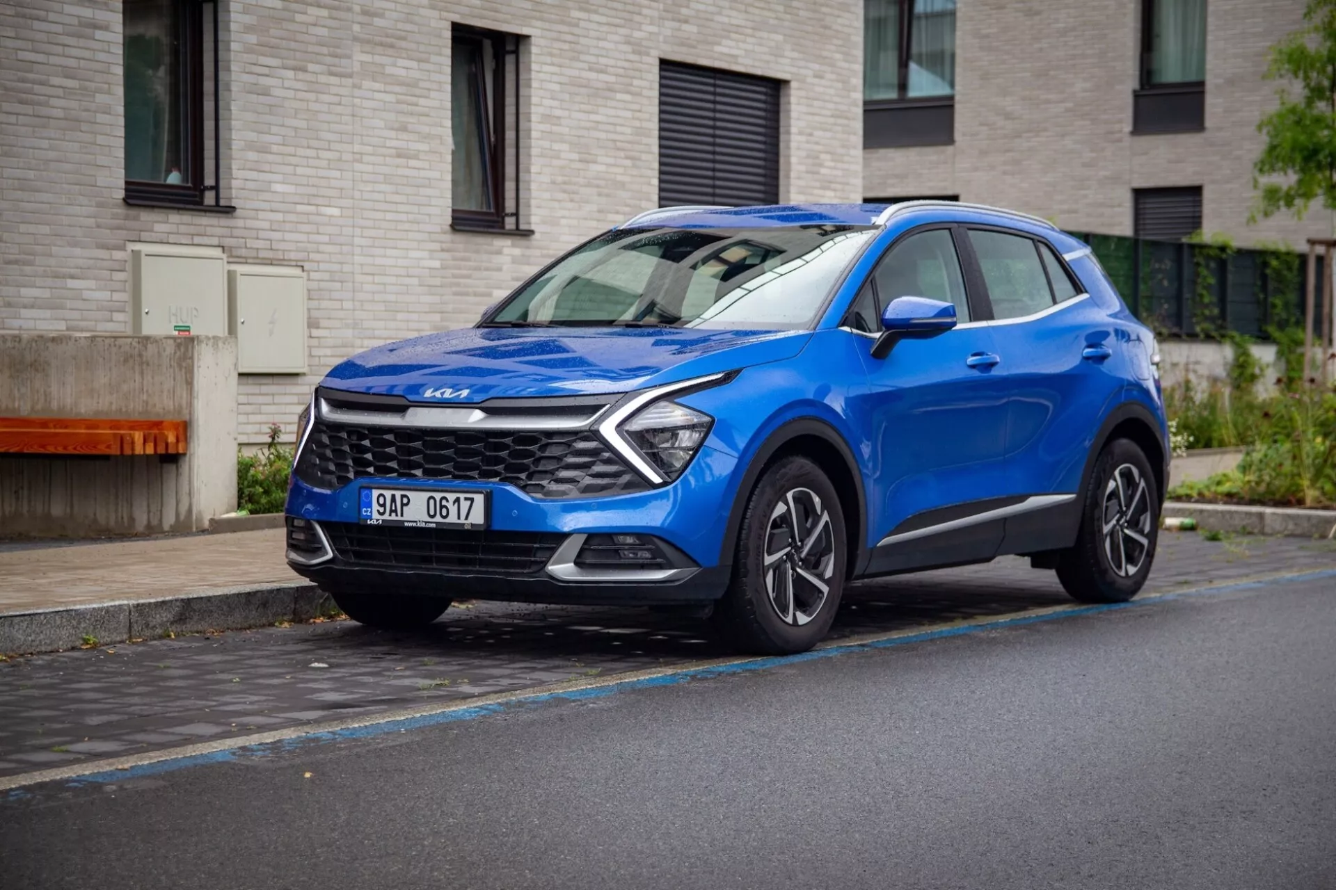 SUV crossover compact Kia Sportage 2022 en bleu, garé sur une rue mouillée montrant sa vue avant trois quartiers dynamique avec des feux de jour à LED, capot sculpté et roues en alliage.