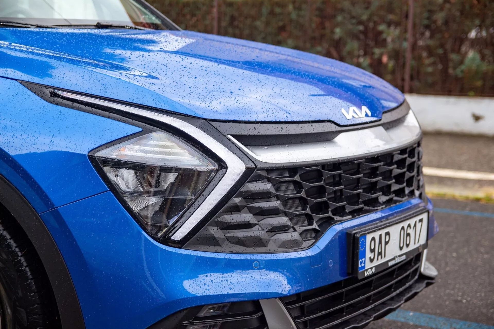 Vue rapprochée d'une Kia Sportage bleue 2022 se concentrant sur le quart avant gauche sous des conditions humides, mettant en lumière la conception dynamique du capot, les phares LED, la grille en nid d'abeille et la technologie des capteurs avancée