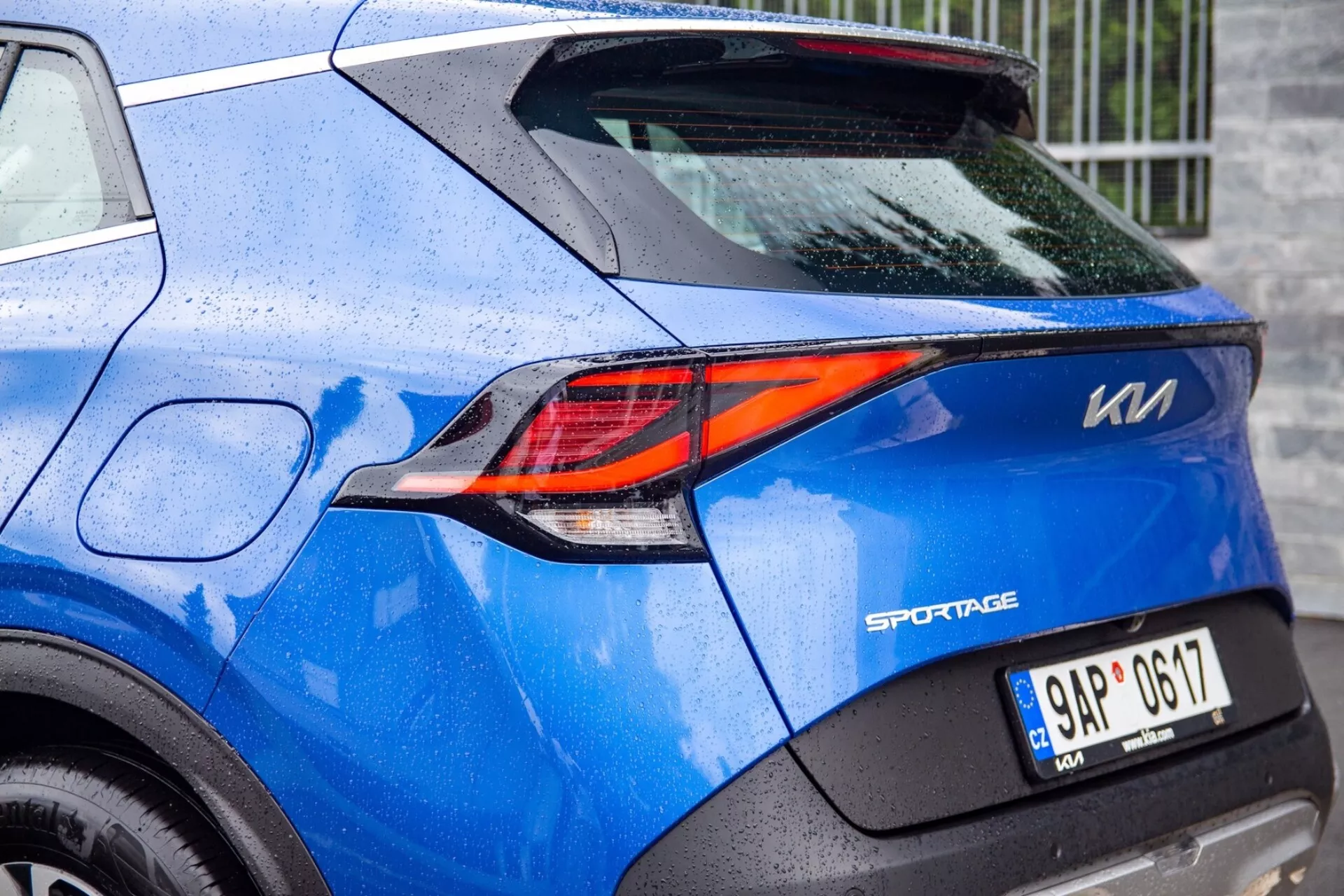 Vue partielle d'un Kia Sportage bleu 2022 mettant en évidence le feu arrière LED en forme de Z sophistiqué et les détails de conception arrière, y compris le badge du modèle, la plaque d'immatriculation et la peinture parsemée de gouttes de pluie