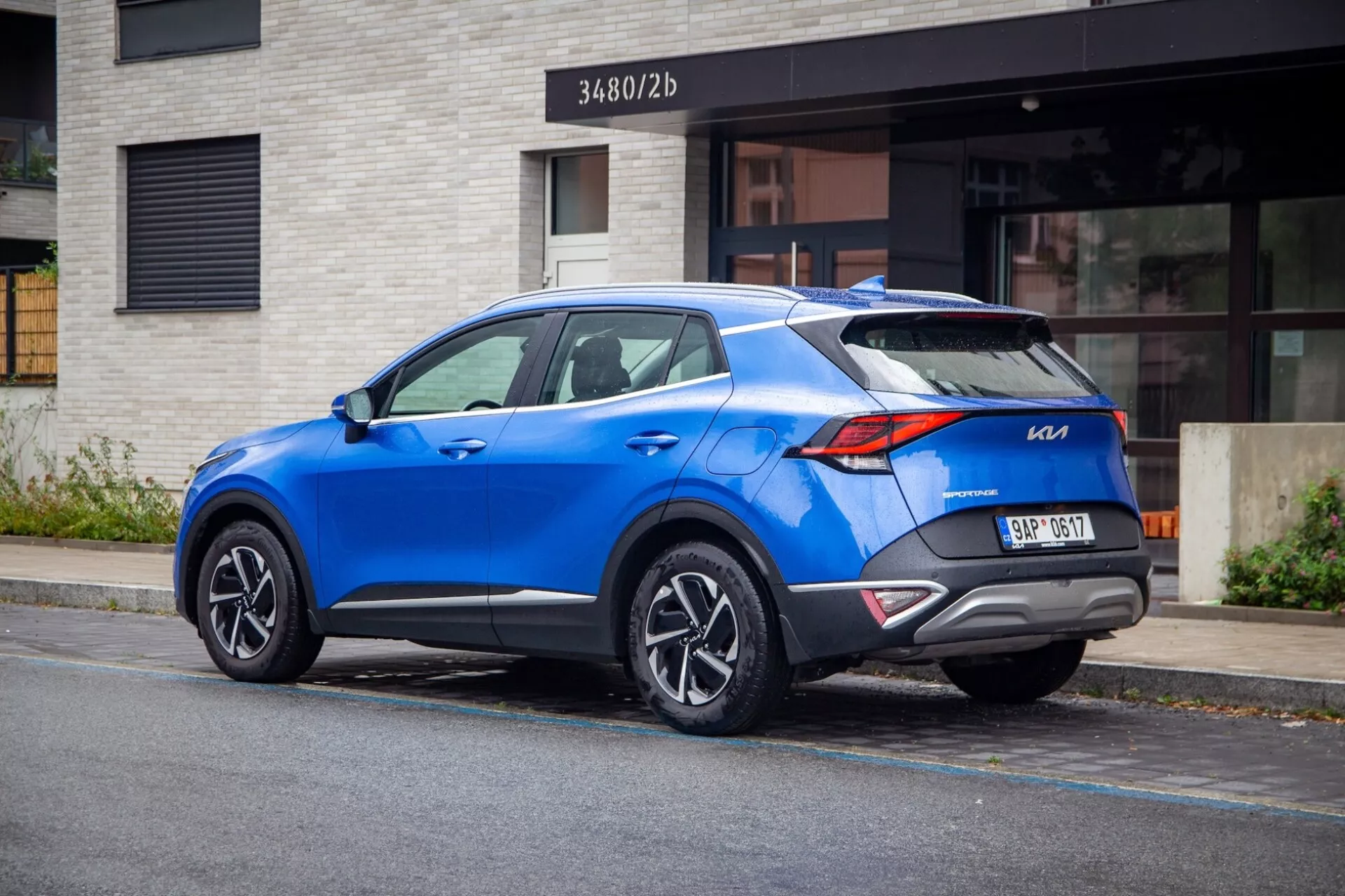 SUV crossover moderne Kia Sportage 2022 garé dans une rue de la ville, montrant la vue arrière trois quarts avec des feux arrière distinctifs, un spoiler de toit sportif, des jantes en alliage à plusieurs branches, une couleur bleue vibrante et des fonctionnalités technologiques modernes potentielles.