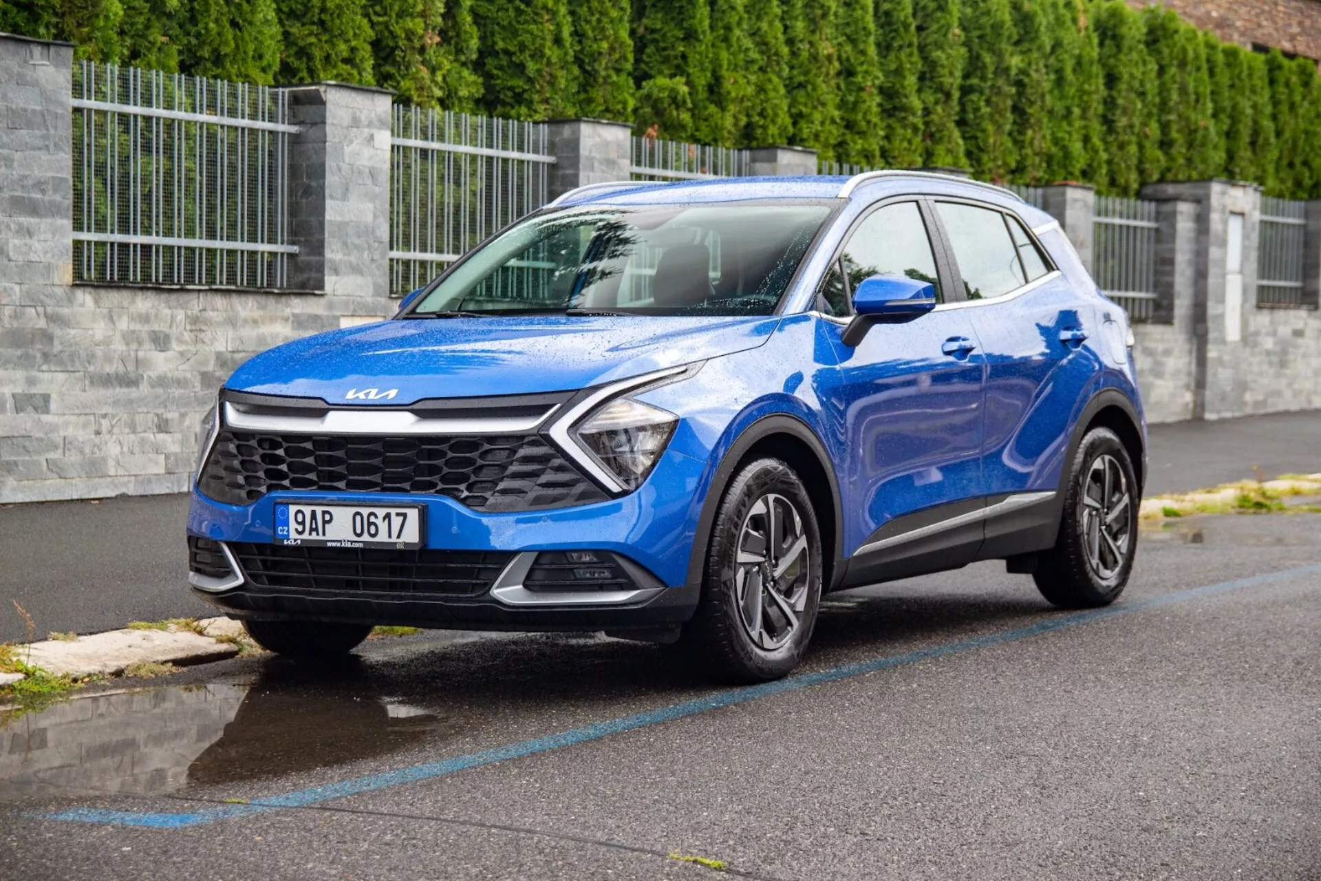 SUV compact Kia Sportage 2022 en bleu garé sur une rue en asphalte mouillé présentant la grille avant, les phares, le pare-chocs, le capot et le profil côté conducteur depuis un angle avant de trois quarts avec une perspective basse pour mettre en évidence la conception du véhicule et les caractéristiques technologiques
