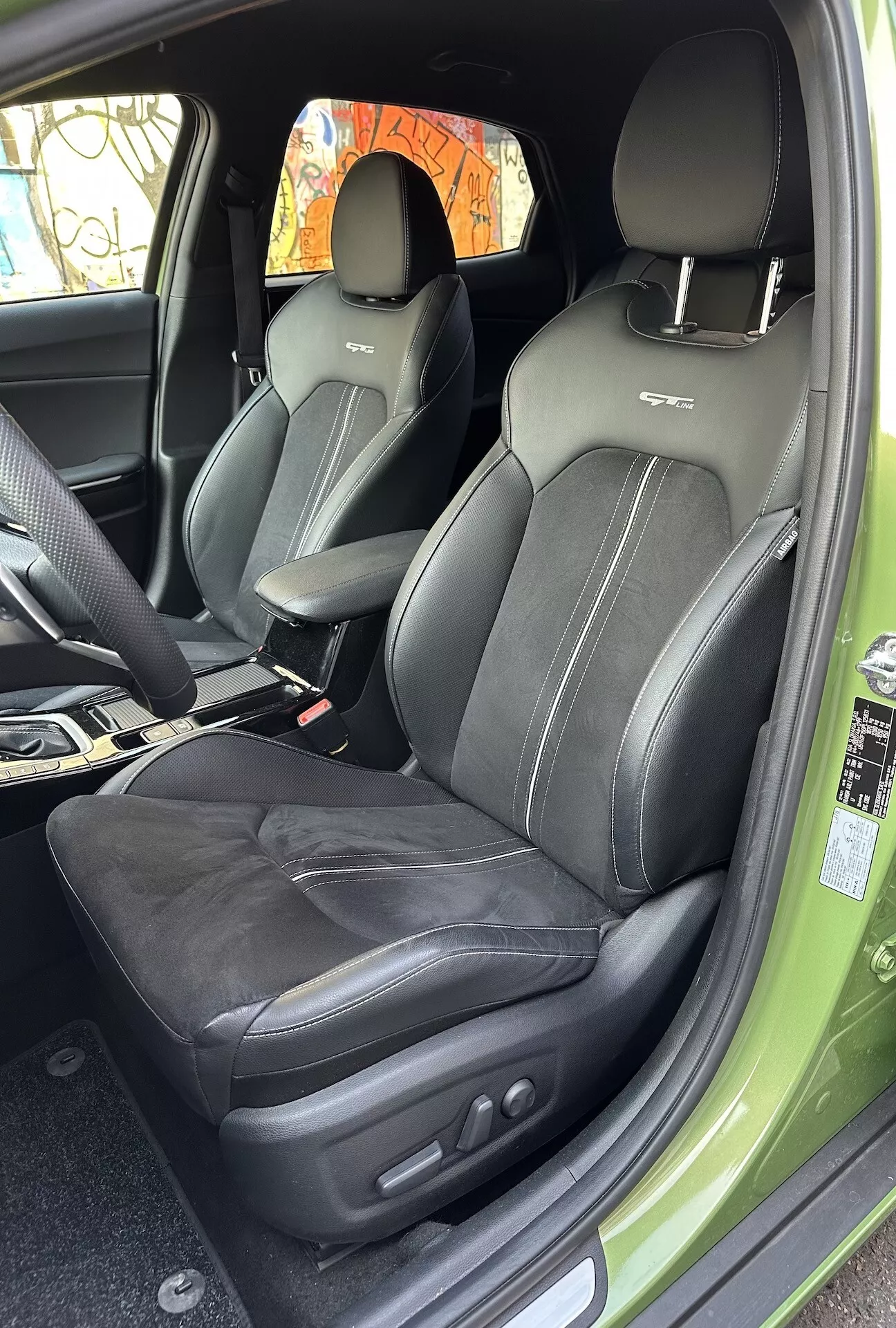 Vue intérieure du Kia XCeed 2022 se concentrant sur le compartiment du passager avant avec revêtement en cuir, coutures complexes et commandes de réglage de siège électroniques, depuis un angle de porte ouverte offrant un aperçu de l'extérieur vert vibrant.
