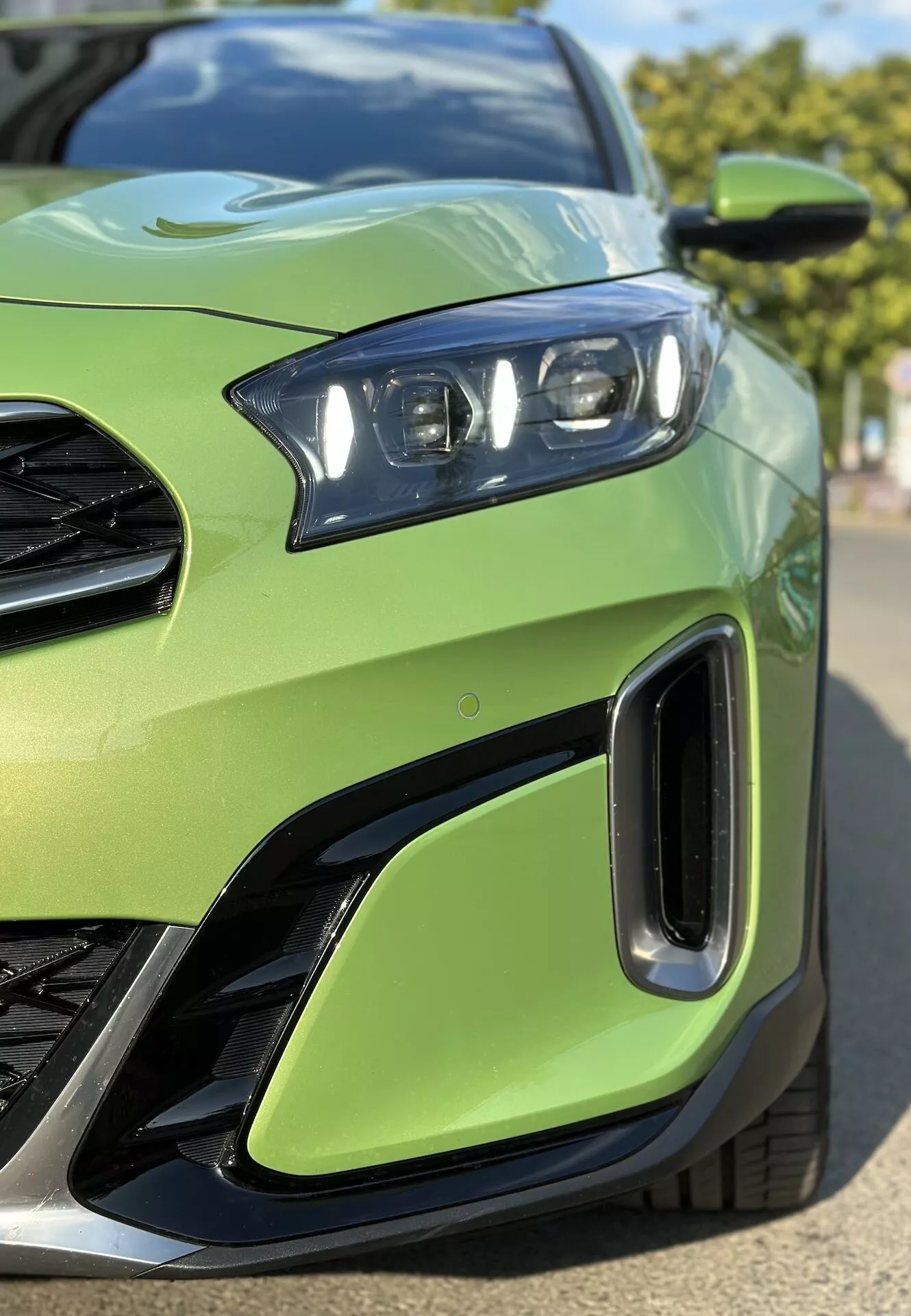 Gros plan sur le quart avant gauche de la Kia Xceed 2022 montrant en détail l'ensemble phare LED, une partie de la grille, les entrées d'air sur le pare-chocs avant, sur une couleur de carrosserie vert lime, soulignant le design sportif du véhicule et la technologie d'éclairage avancée.