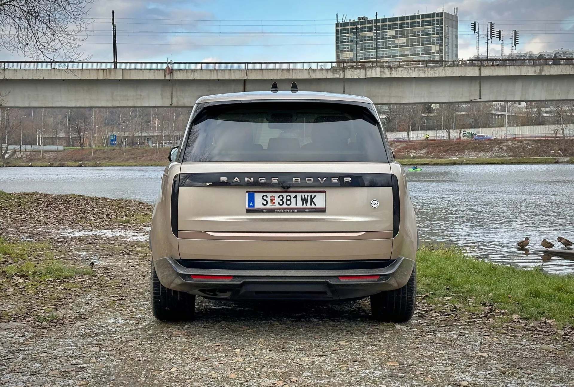 Vue arrière du Range Rover 2021 avec angle de vue direct à hauteur des yeux, stationné près d'un plan d'eau avec des canards et un viaduc en arrière-plan, présentant le nom du modèle, les feux arrière, les capteurs de stationnement et le système d'échappement double en beige mat