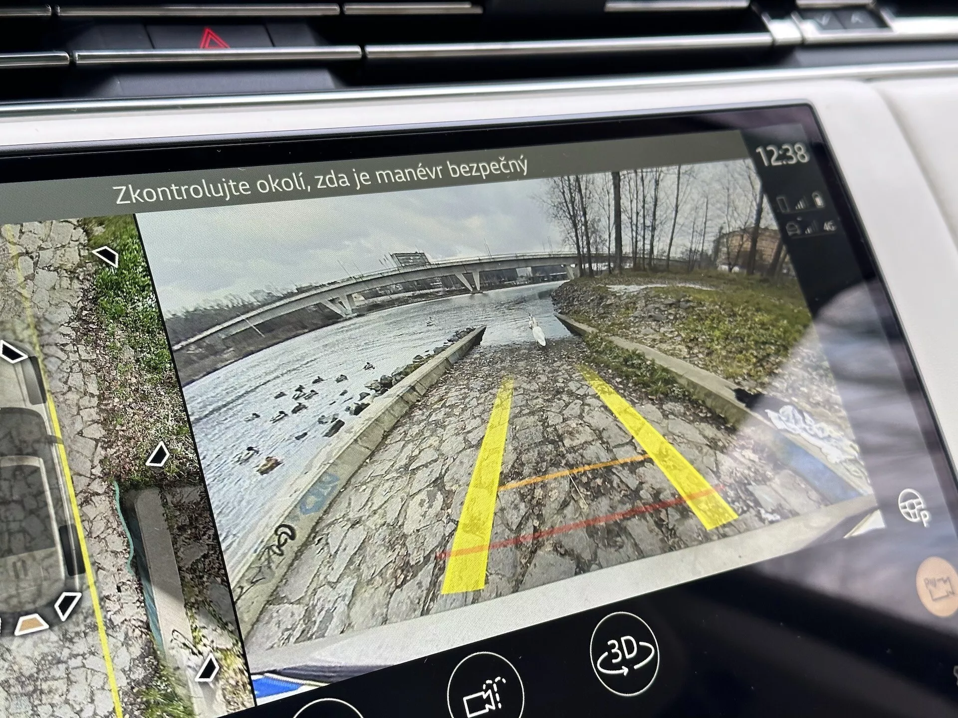 Affichage d'infodivertissement du Land Rover Range Rover 2021 montrant la vue arrière de la caméra de recul avec des lignes de guidage dynamiques dans un espace de stationnement près d'un plan d'eau, détaillé avec de la végétation, des icônes d'aide au stationnement et un texte de rappel de sécurité