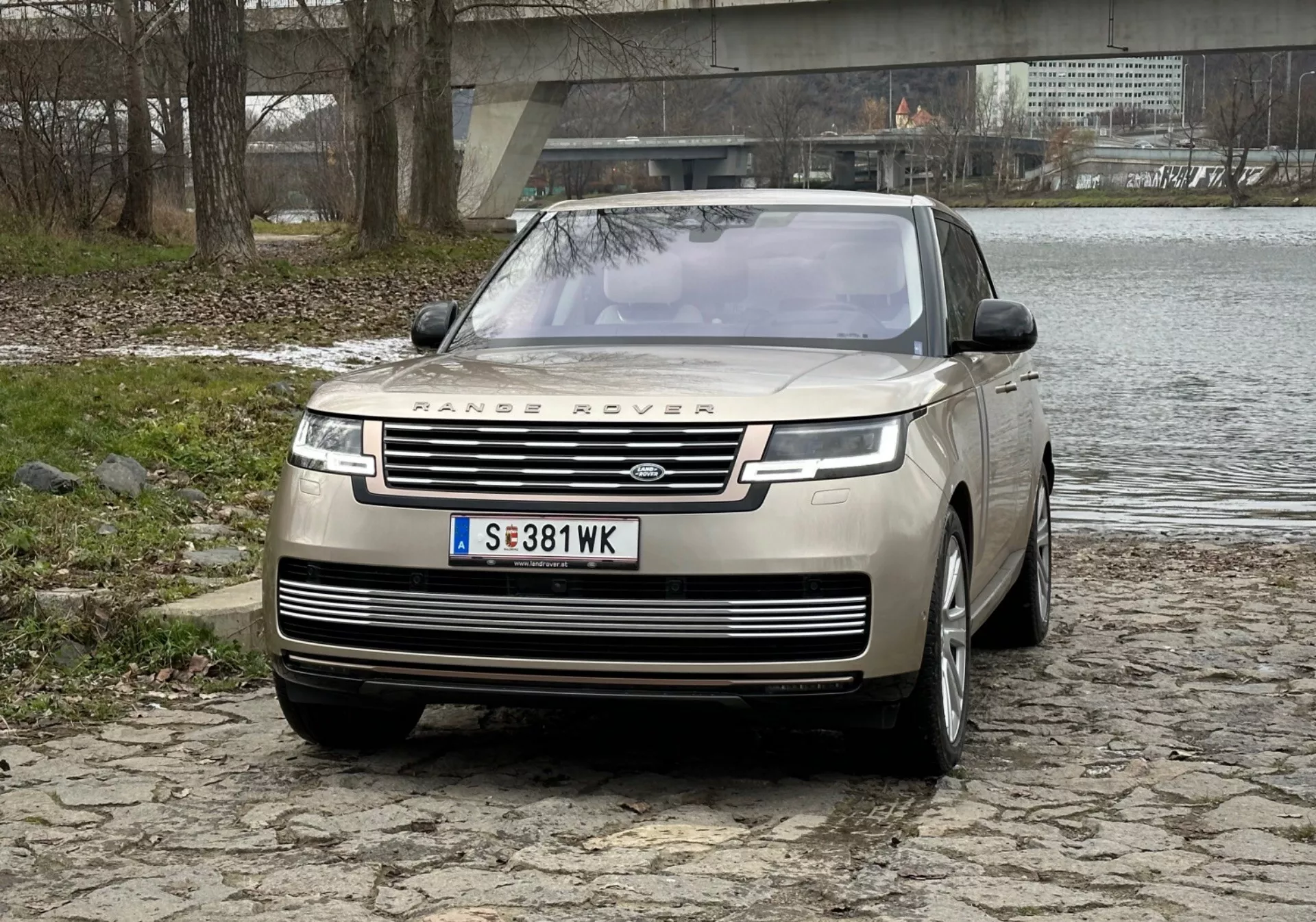 SUV de luxe Range Rover 2021 en argent, présentant une grille avant proéminente avec marquage, feux de jour LED élégants, pare-chocs avant avec capteurs de stationnement et design de capot clamshell sur une surface en pavés près d'une rivière