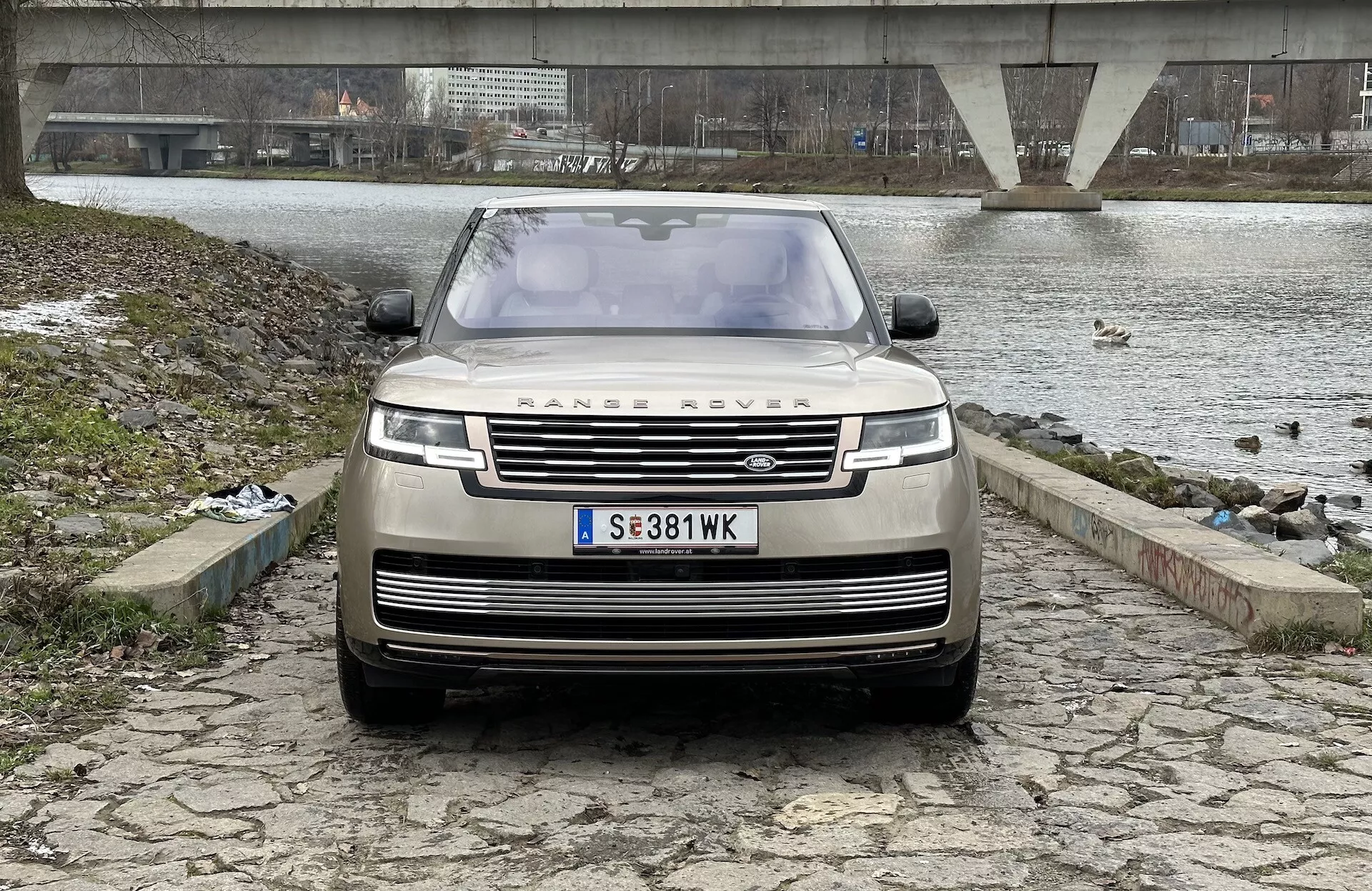 Land Rover Range Rover 2021 photographié depuis un angle frontal et légèrement bas, mettant en évidence la grille, le pare-chocs avant et le capot, avec l'emblème emblématique RANGE ROVER, des phares LED modernes et une technologie avancée d'aide à la conduite, sur fond de cours d'eau avec un pont et un ciel couvert