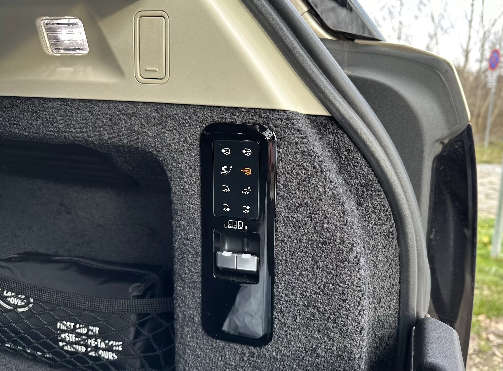 Vue rapprochée du panneau de contrôle électronique du coffre à l'intérieur d'une Land Rover Range Rover 2021, montrant des boutons pour des opérations comme l'ouverture et la fermeture électroniques, le contrôle du couvercle de charge, potentiellement une lumière de coffre ou une prise de courant, des réglages des sièges arrières depuis le coffre et le déclenchement d'urgence, situé contre la paroi latérale du coffre tapissée et le plafond de couleur claire avec fixture de lumière de chargement.