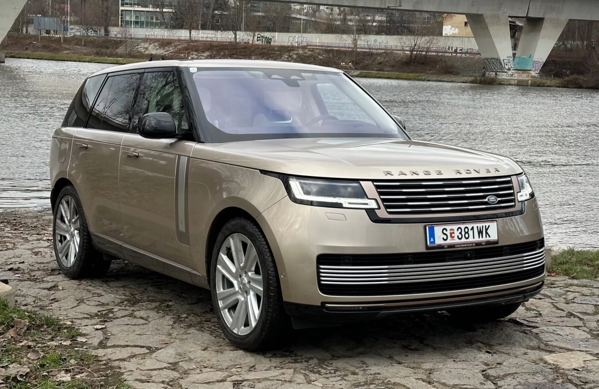 SUV de luxe Land Rover Range Rover 2021 garé au bord d'une rivière avec un pont en arrière-plan, présentant son design moderne et ses caractéristiques technologiques, y compris des phares LED, une calandre avant avec emblème, des jantes en alliage à plusieurs branches et un toit panoramique.