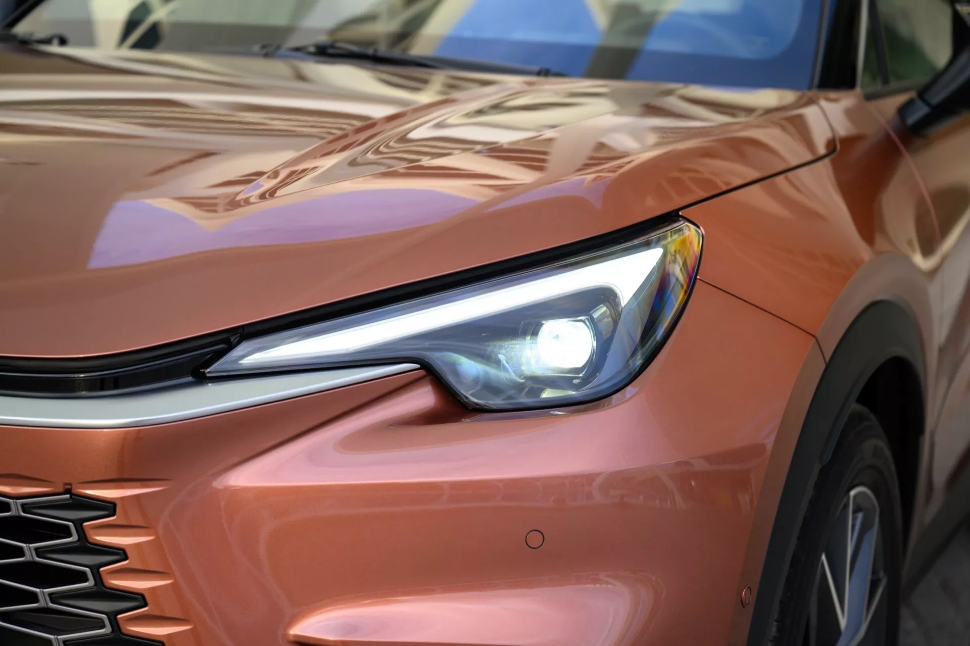 Image en gros plan du quart avant gauche d'une Lexus LBX 2024, première génération, mettant en évidence le phare LED, la grille et le capot ainsi que l'aile profilés depuis un angle bas, soulignant son design épuré et ses éléments technologiques modernes.