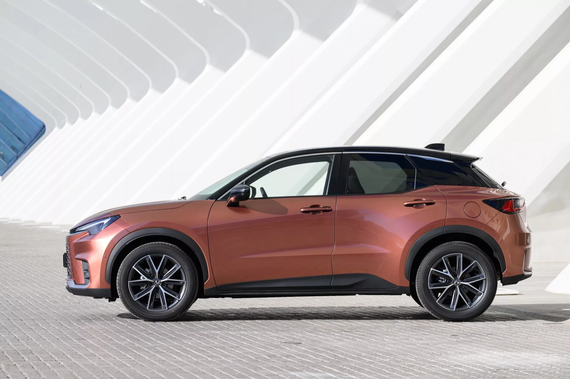 Profil latéral du Lexus LBX 2024 sous éclairage artificiel, mettant en avant la couleur cuivre métallique, phares LED élégants et structure géométrique en arrière-plan