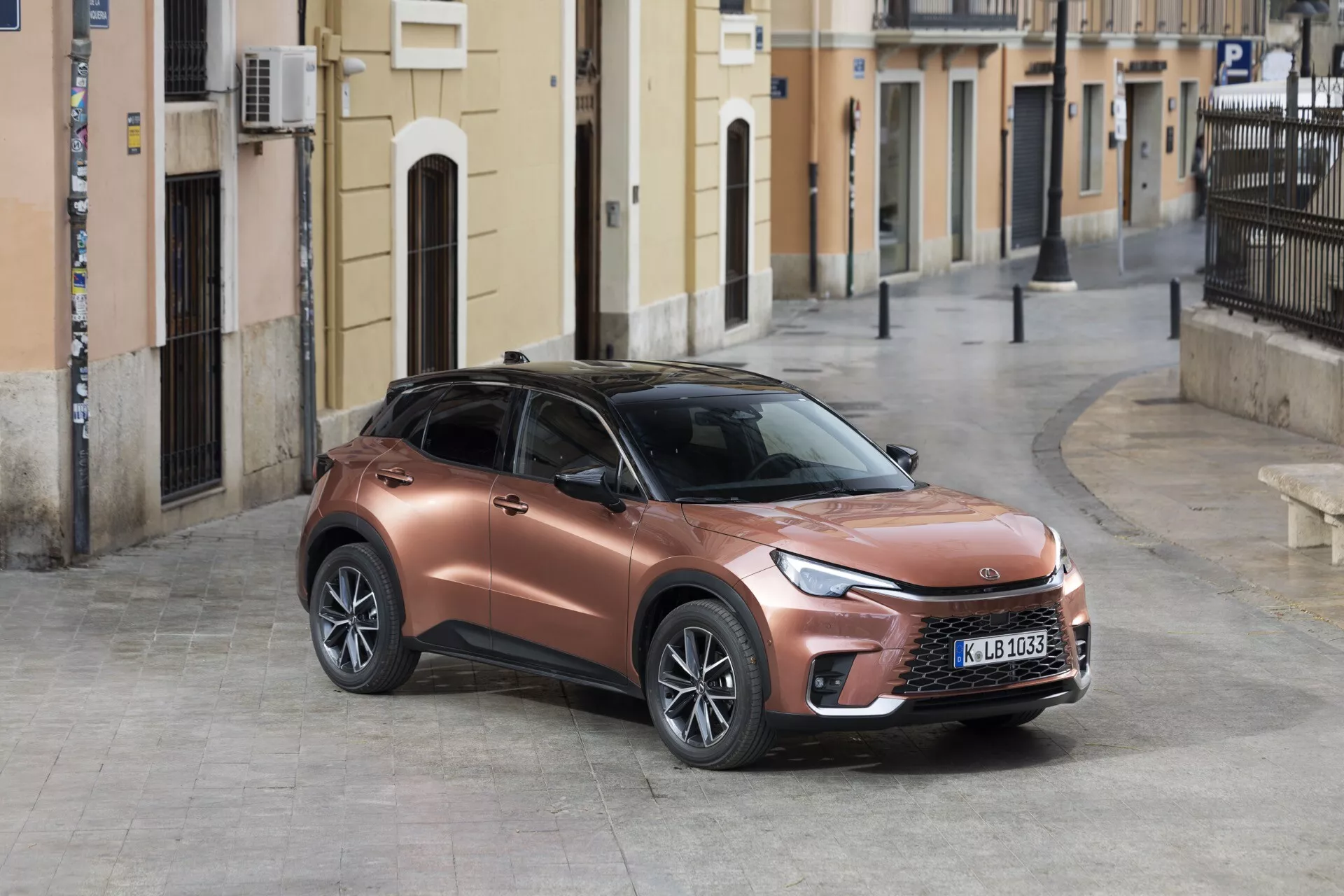 SUV crossover moderne Lexus LBX 1ère génération 2024 garé sur une rue pavée avec une vue de trois quarts du côté avant gauche mettant en vedette sa finition métallique bronze, des phares LED élégants et un design sportif surélevé