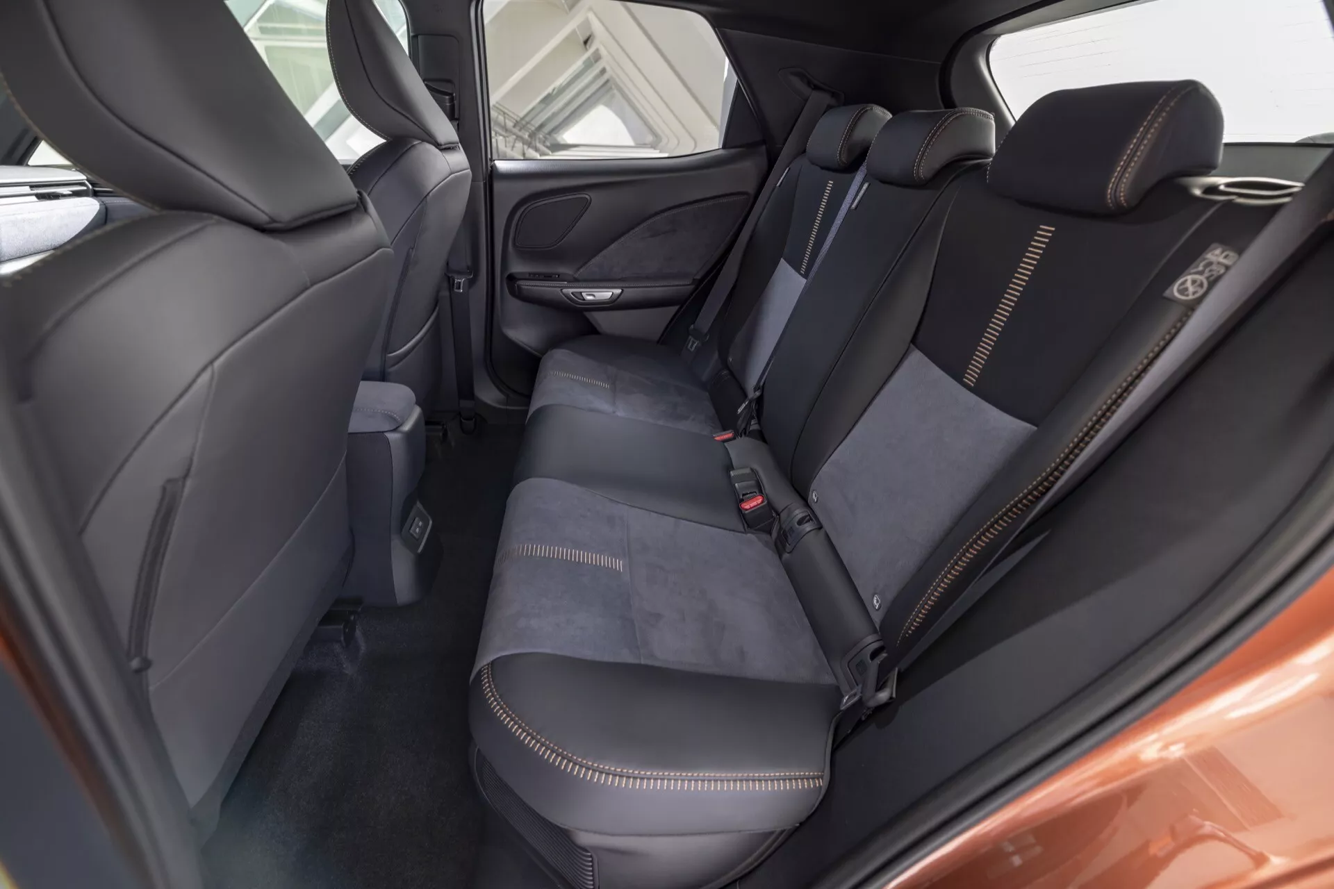 Vue intérieure de la Lexus LBX 1ère génération 2024 depuis le compartiment passager arrière montrant la zone du siège arrière, détails de l'upholsterie avec couture contrastante blanche, dossiers matelassés, accoudoir rabattable, grille du haut-parleur sur la porte et poche de rangement au dos du siège avant