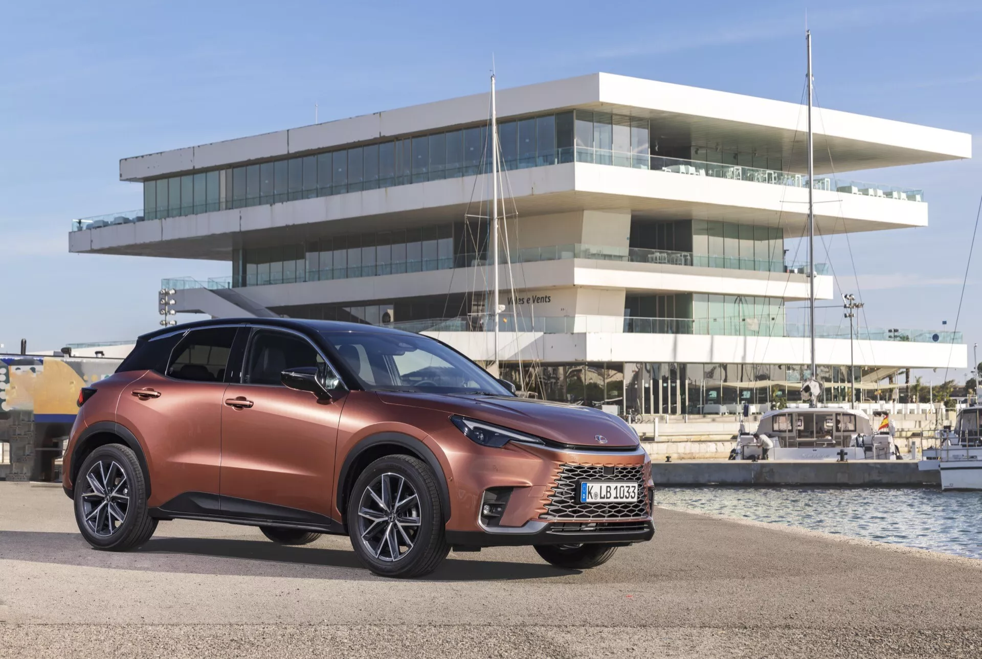 SUV crossover moderne Lexus LBX 1ère génération 2024 en cuivre métallique, présentant grille avant, phares LED et roues en alliage, positionné en extérieur avec une marina et un bâtiment contemporain en arrière-plan, soulignant son orientation vers un style de vie urbain/côtier.