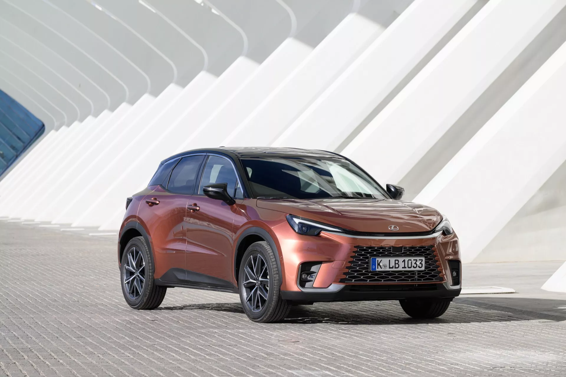 Lexus LBX 1ère génération 2024 présentée en pleine lumière, dotée d'une grille avant distinctive et de phares LED élégants, sur fond d'architecture moderne aux formes géométriques angulaires. L'angle met en valeur la carrosserie aérodynamique de la voiture, la peinture métallisée et les roues en alliage, soulignant son design de SUV crossover et l'ingénierie automobile avancée.