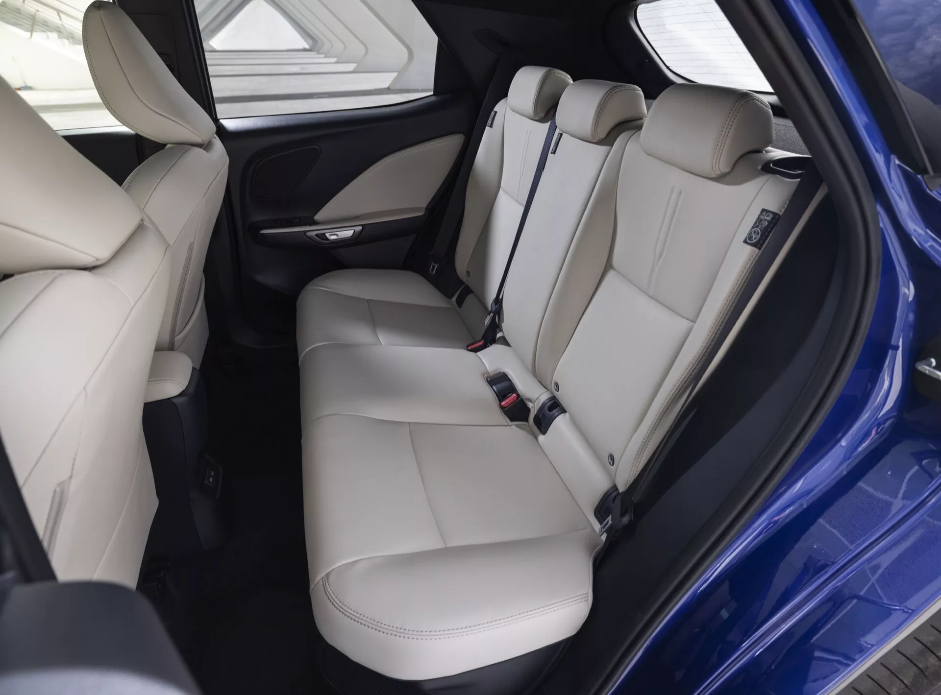 Intérieur du Lexus LBX 1ère Génération 2024 avec sièges arrières en cuir beige clair premium avec appuis-tête, ceintures de sécurité, panneau de porte avec commandes, section arrière de la console centrale comprenant des bouches d'air et des ports de chargement, dans des conditions de lumière optimales mettant en évidence les caractéristiques de confort et de sécurité