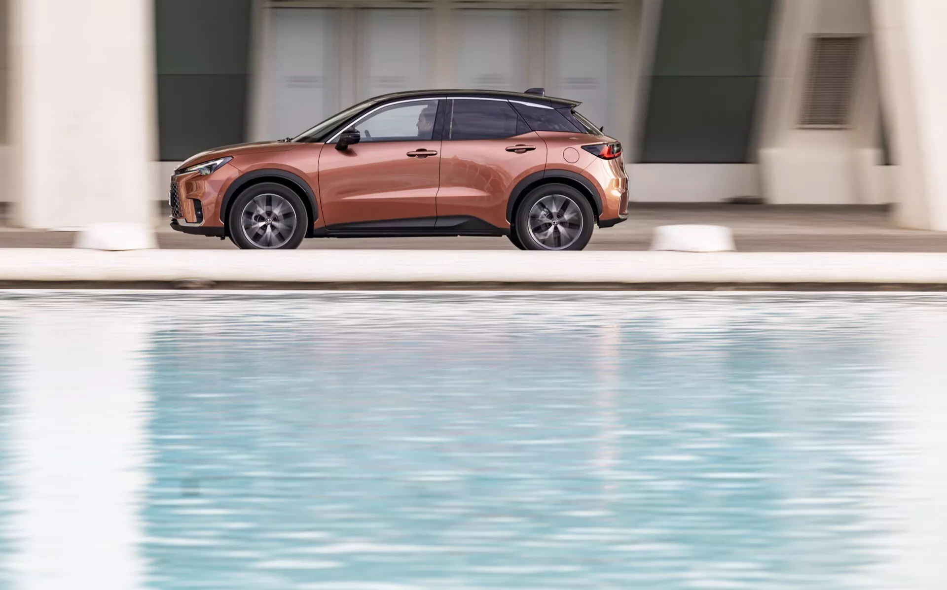 Vue de profil latéral de la Lexus LBX 1ère génération 2024 présentant des jantes en alliage, un design à quatre portes, des rétroviseurs latéraux avec clignotants intégrés, des vitres teintées, des barres de toit et des détails de garniture, sur un fond serein avec un plan d'eau réfléchissant