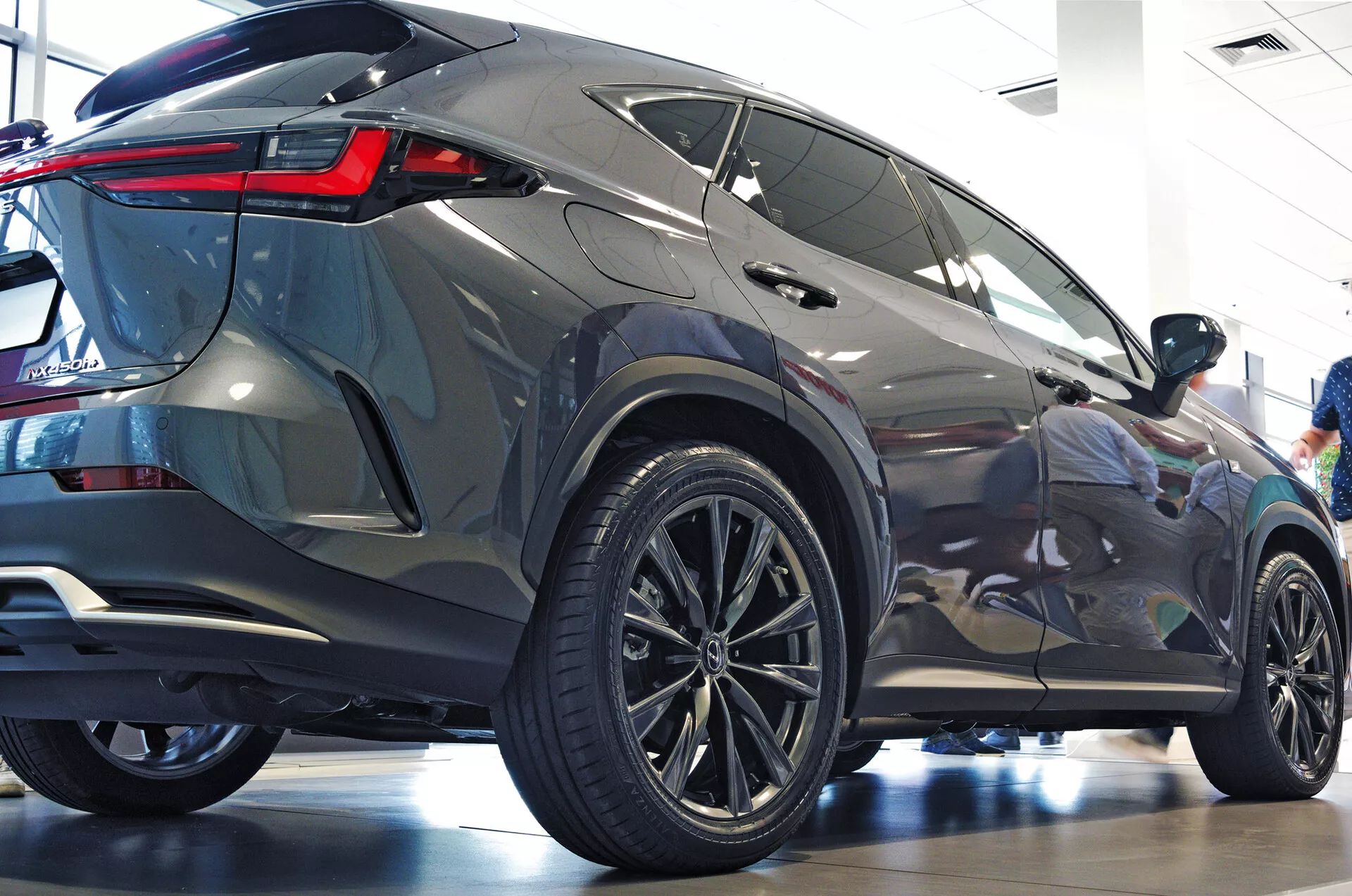 Angle de trois quarts arrière du SUV Lexus NX 2021 montrant le profil latéral et postérieur avec de grandes roues en alliage multi-branchures, des pneus à profil bas, des feux arrière LED angulaires et vifs, une carrosserie métallique, des vitres teintées, un spoiler arrière intégré et un environnement de showroom poli mettant en avant l'esthétique moderne et les caractéristiques de luxe