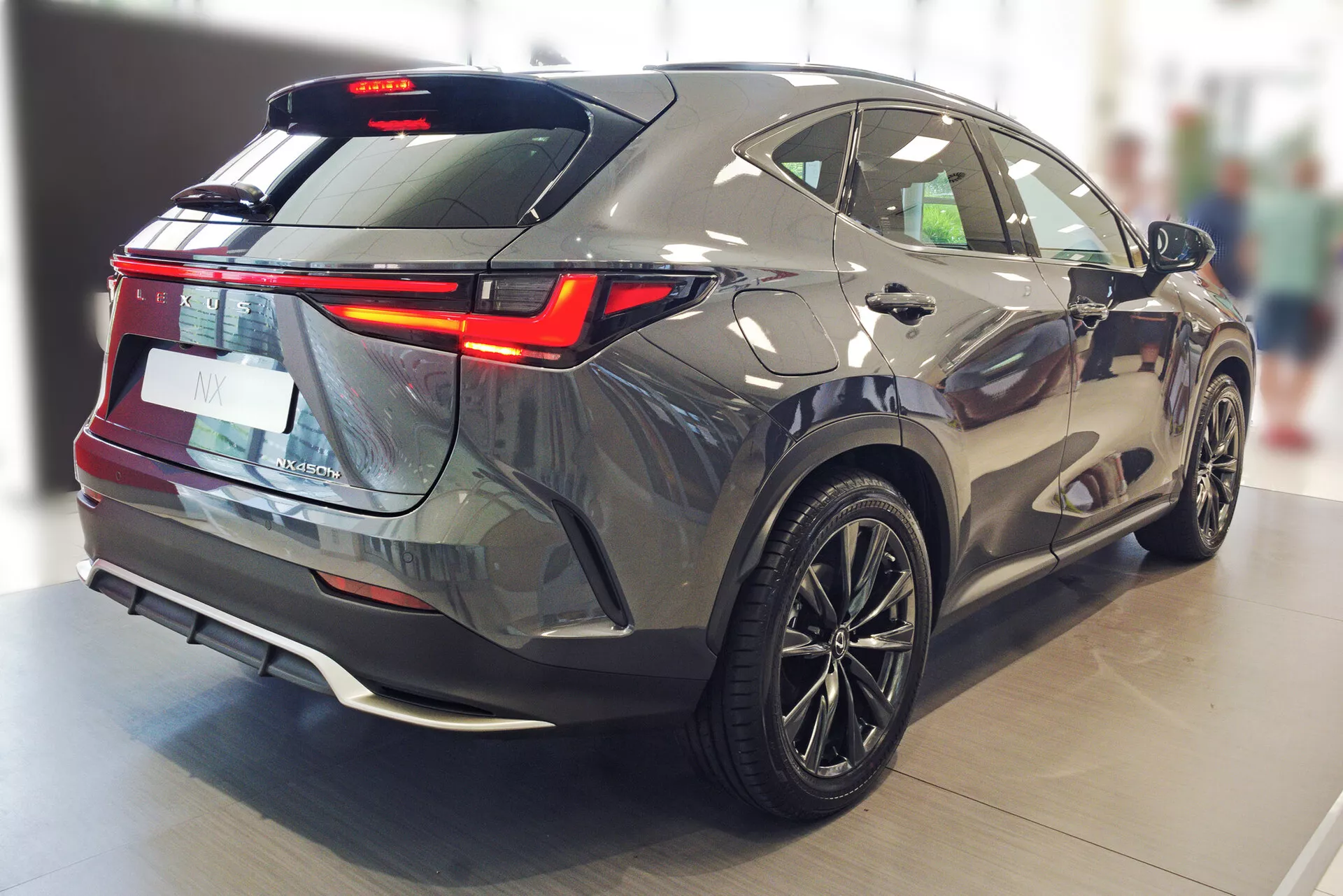 Image capturant la Lexus NX 2021 sous un angle arrière trois-quarts, mettant en évidence ses contours aérodynamiques et élégants, les feux arrière à LED, la vitre arrière fortement inclinée, le spoiler arrière, l'essuie-glace arrière, le pare-chocs arrière bicolore avec capteurs de stationnement, l'élément de style diffuseur, les jantes en alliage multi-branches et sa couleur grise métallisée sur le sol poli d'un showroom intérieur.