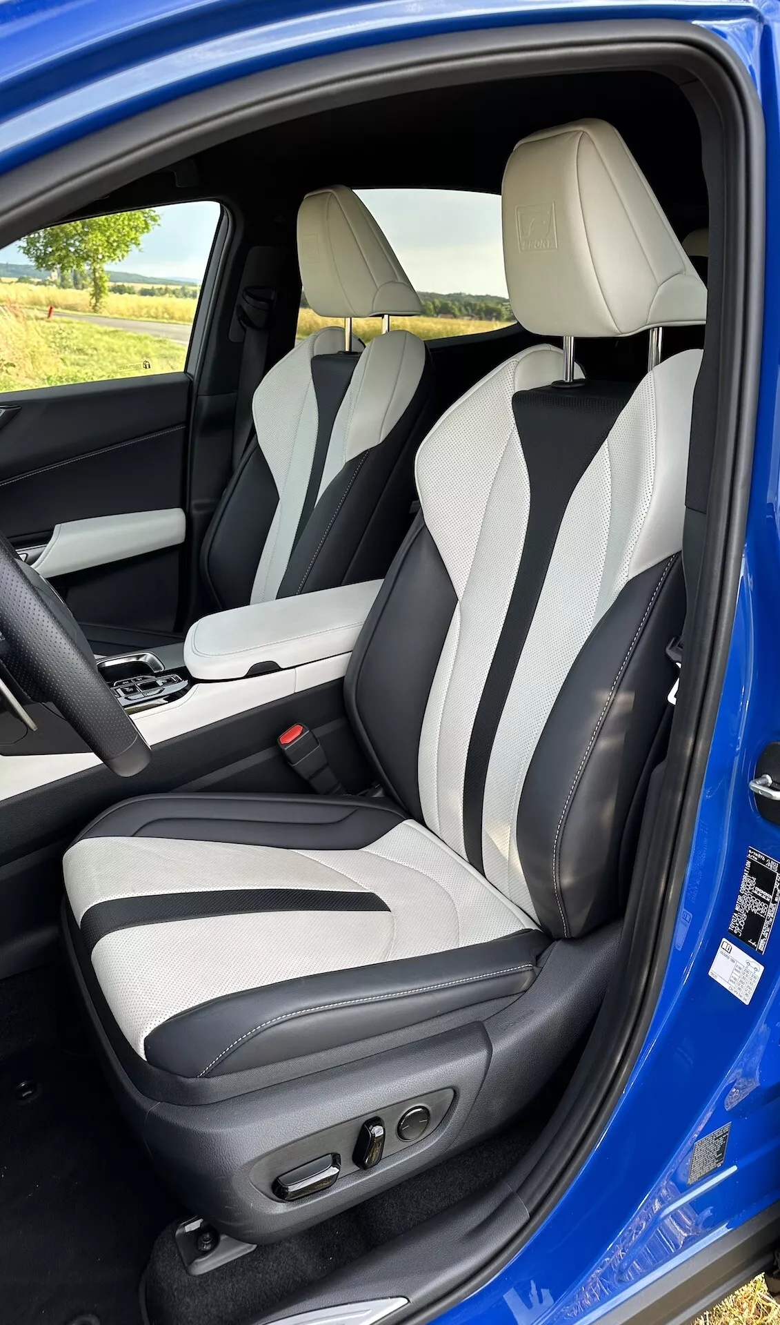 Vue intérieure à travers la porte ouverte du côté conducteur d'une Lexus NX 2021, montrant la zone du conducteur depuis une légère élévation, avec une sellerie en cuir bicolore, des commandes avancées de siège conducteur, des renforts latéraux, des appuie-têtes intégrés et une console centrale avec levier de vitesse automatique