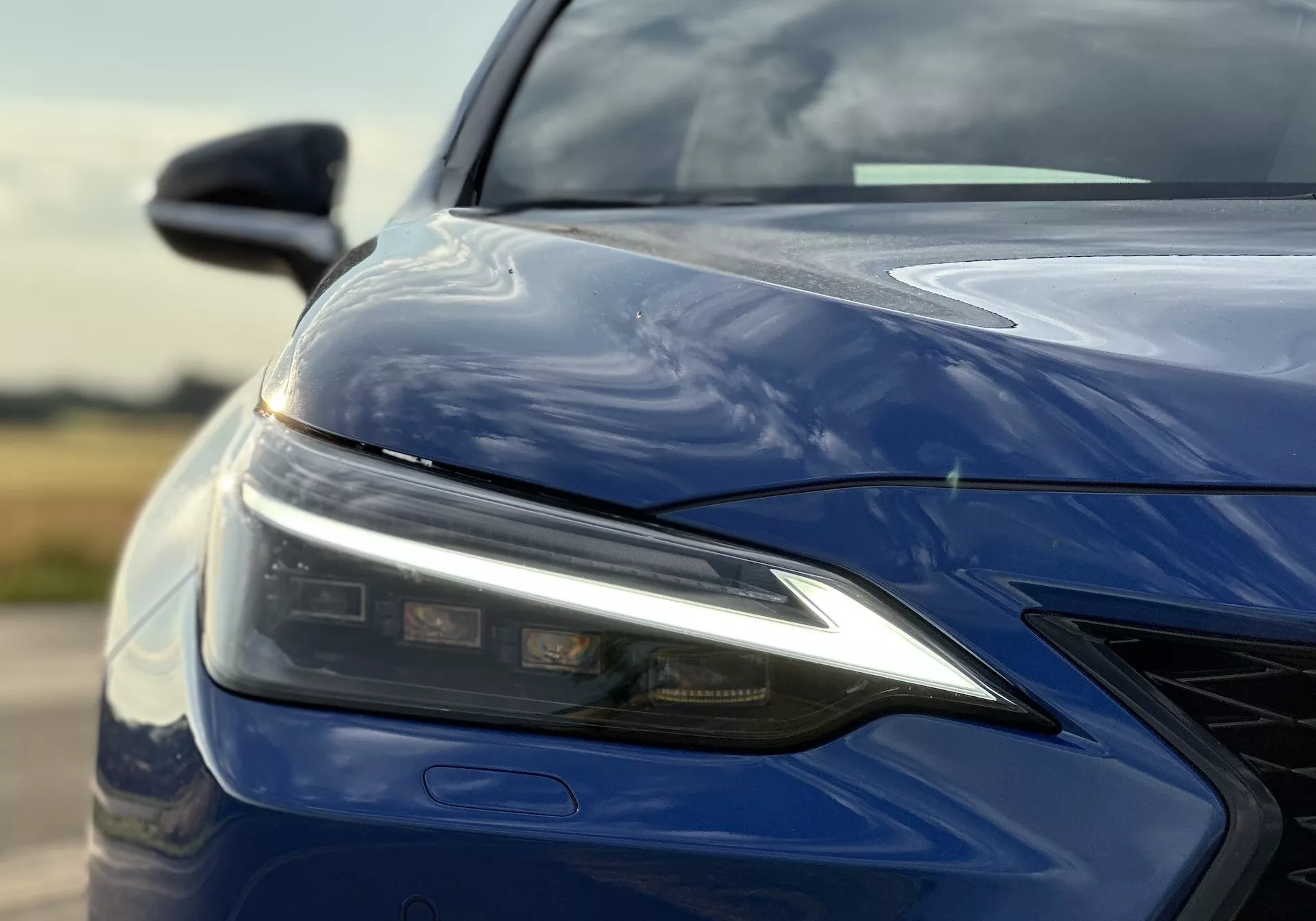 Gros plan sur le quart avant gauche du Lexus NX 2021 avec assemblage de phare LED, capot sculpté, pare-choc avant avec grille et finition bleu profond brillant, mettant en évidence la technologie d'éclairage avancée et la conception aérodynamique.