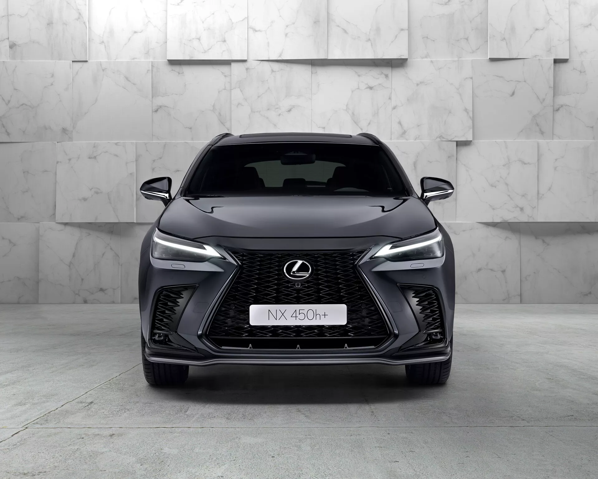 Vue frontale du Lexus NX 450h+ 2021, un SUV moderne et élégant mettant en avant sa grille en forme de fuseau avec un motif en maille, des phares LED étroits, le badge Lexus chromé avec contour bleu et un design agressif du pare-chocs avant. Le caractère hybride de la voiture est indiqué par le badge 'h+' sur la grille. L'environnement du studio met en valeur les courbes et les caractéristiques du véhicule avec un éclairage propre et contrôlé.