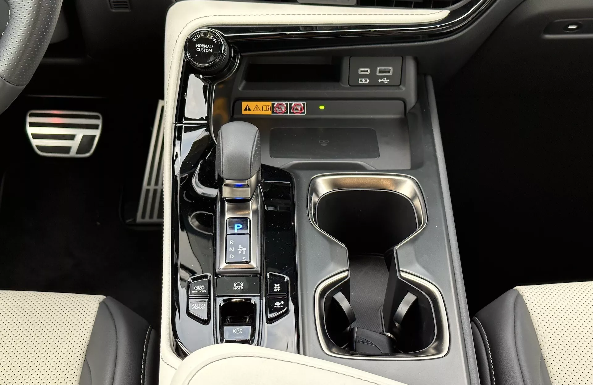 Vue de la console centrale du Lexus NX 2021 montrant le levier de vitesse automatique avec soufflet en cuir, le sélecteur de mode de conduite, les porte-gobelets, les boutons de commande du véhicule, les éventuels raccourcis vers le système d'infodivertissement et les éléments de design intérieur de luxe