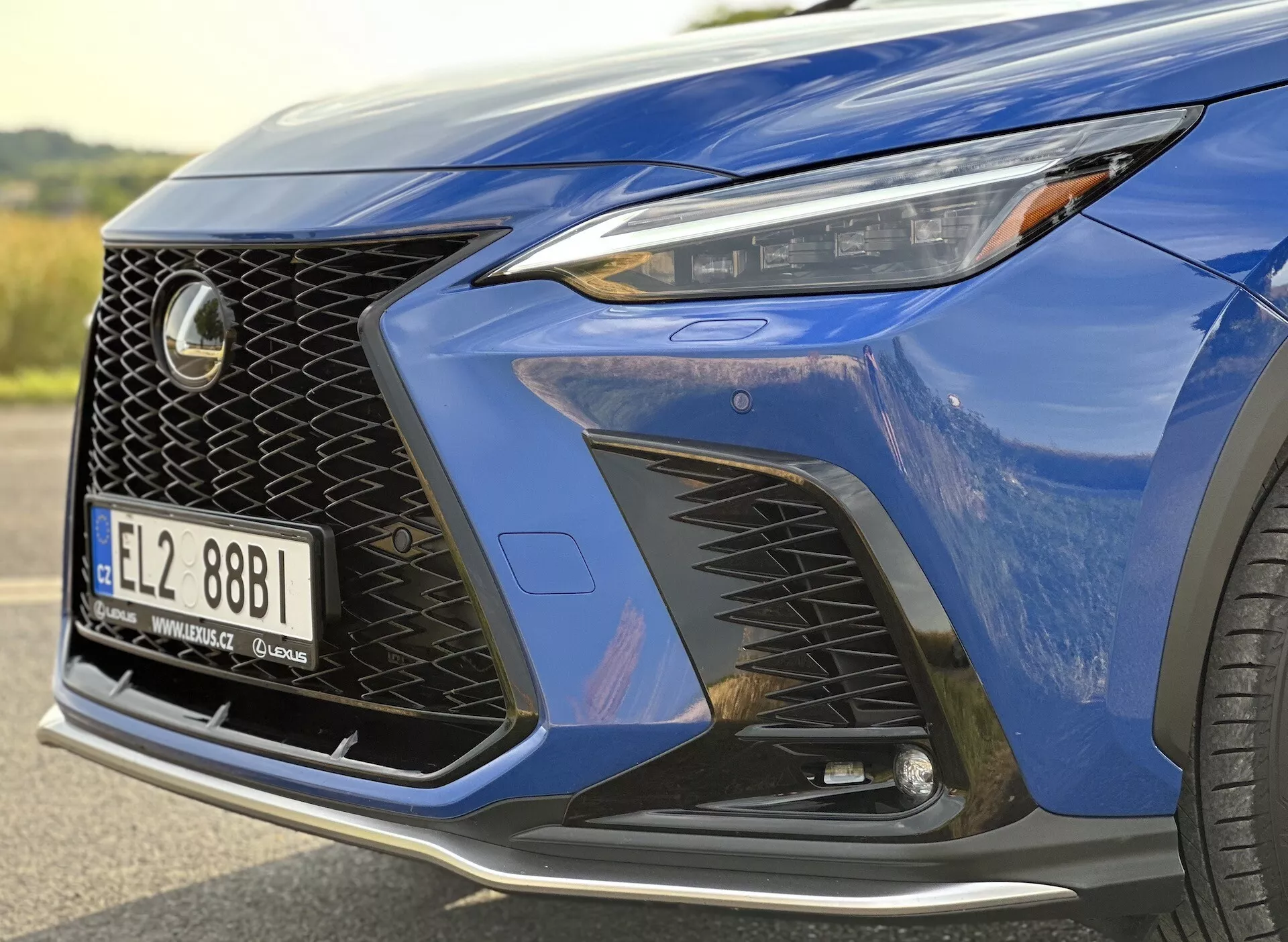 Gros plan sur le coin avant du Lexus NX 2021 mettant en vedette le phare avec technologie LED, design distinctif de la grille en maille et pare-chocs avant aérodynamique avec prises d'air, souligné par une peinture métallisée bleu profond et un aperçu d'une roue en alliage de luxe.
