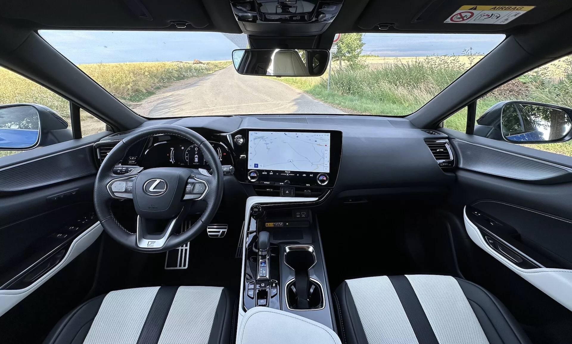 Vue intérieure du Lexus NX 2021 du point de vue du conducteur, montrant un volant centré avec le logo Lexus et des boutons de contrôle, un grand écran tactile d'infodivertissement sur le tableau de bord, des commandes climatiques minimalistes, un sélecteur de vitesse automatique sur la console centrale avec porte-gobelets et rangement, des sièges en matériel bicolore, et une vue partielle sur un paysage rural à travers le pare-brise.