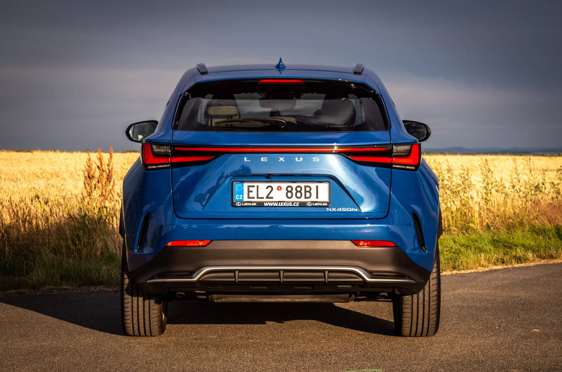 Vue arrière du SUV bleu Lexus NX 2021 sur route en asphalte avec champ doré en arrière-plan, mettant en avant la technologie hybride et les caractéristiques de design