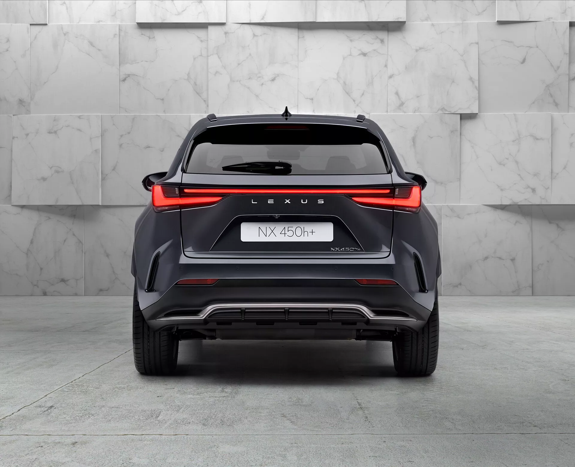 Vue arrière du SUV hybride Lexus NX 450h, capturée sous un angle bas légèrement incliné vers la gauche, mettant en avant la conception des feux arrière sur toute la largeur, le logo 'LEXUS', la désignation du modèle 'NX 450h', le nom 'NEXUS' et le symbole hybride 'h'. Le pare-chocs avec ses éléments de style angulaire, ses caractéristiques ventilées et une plaque de protection argentée, avec une pointe d'échappement double visible du côté gauche, le tout dans un environnement de showroom avec un sol brillant et un fond de ton neutre.