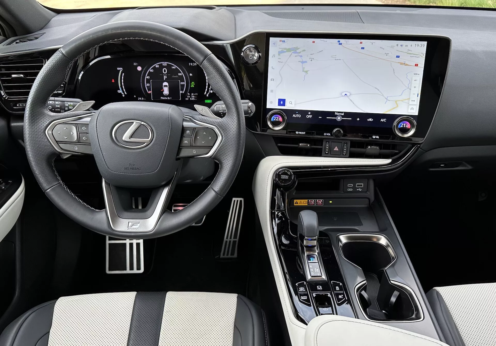 Vue intérieure du Lexus NX 2021 montrant la zone du conducteur et la console centrale sous un angle élevé, avec le logo Lexus sur le volant doté de commandes, un tableau de bord numérique éclairé, un grand système d'infodivertissement tactile avec navigation GPS, boutons de contrôle du climat et de l'audio, sélecteur de vitesse ergonomique, commandes de mode de conduite, matériaux de luxe sur les sièges et points de contact, accents métalliques, et porte-gobelets visibles et compartiment de rangement.