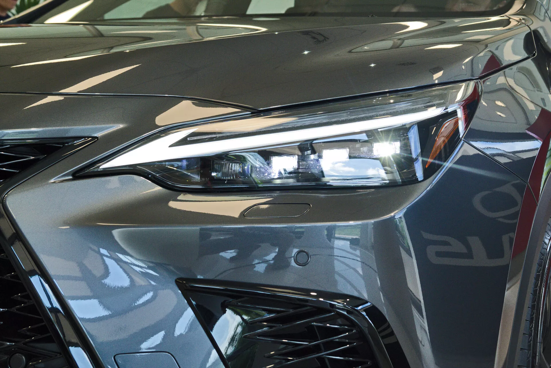Vue rapprochée de l'avant d'une Lexus NX 2021, se concentrant sur l'ensemble du phare côté conducteur avec une technologie d'éclairage avancée à LED et une partie de la grille au style agressif. L'image capture la brillance réfléchissante de la peinture métallique argentée et les lignes dynamiques contribuant à l'esthétique aérodynamique du véhicule. Photographié en intérieur, probablement dans un showroom, soulignant le design luxueux et la finition de haute qualité du véhicule.