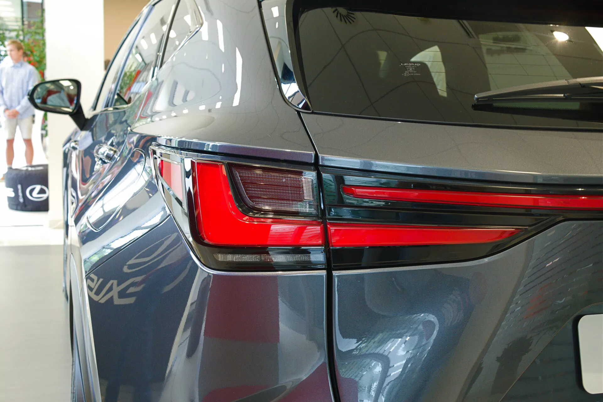 Gros plan sur la Lexus NX 2021 sous un angle arrière trois quarts mettant en évidence le design élégant des feux arrière à LED, une partie du panneau arrière et une portion du couvercle du coffre. L'image montre la peinture sombre et brillante de la voiture, les lignes aérodynamiques du corps et suggère un cadre de showroom intérieur avec des reflets d'un autre véhicule, des éléments architecturaux et une personne marchant en arrière-plan.