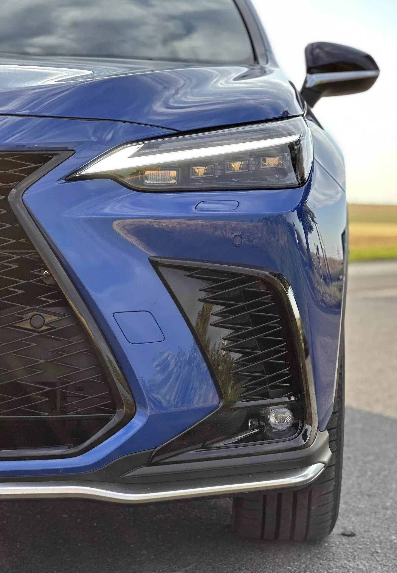Gros plan sur le coin avant gauche d'une Lexus NX 2021 en bleu foncé mettant en lumière la conception avancée des phares LED, le motif complexe de la grille, le pare-chocs avant sportif et les éventuelles caractéristiques adaptatives d'éclairage et aérodynamiques avec une finition métallique brillante
