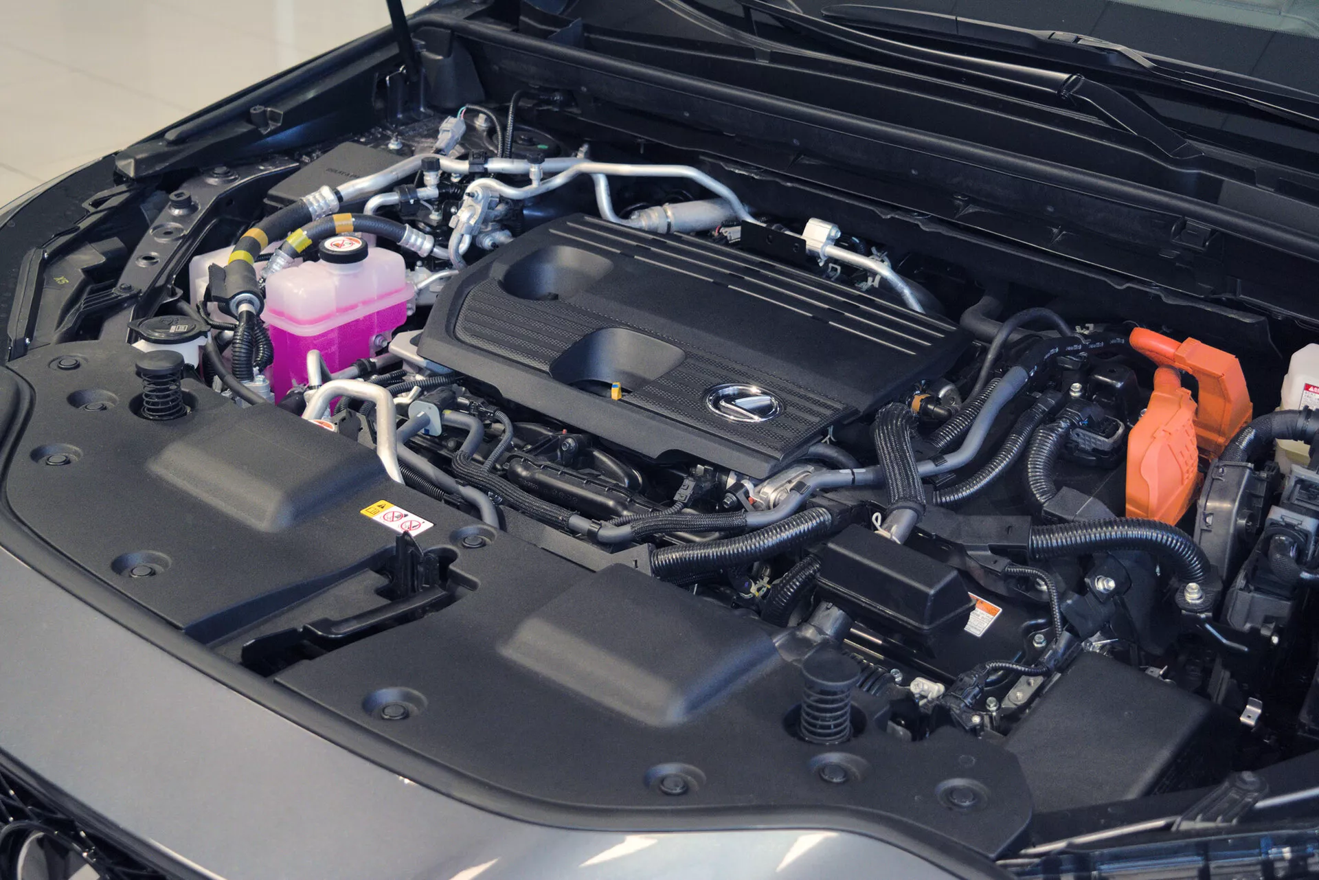 Vue rapprochée du compartiment moteur d'une voiture avec capot ouvert, révélant un compartiment moteur bien organisé et propre. Le cache-moteur arbore un logo bien visible, indiquant la marque, et divers composants tels que des réservoirs colorés, des câbles sécurisés et une batterie visible avec un couvercle de borne positive rouge. L'agencement suggère un véhicule moderne et peut-être luxueux, mettant en avant la complexité de son système électrique et son état général impeccable.
