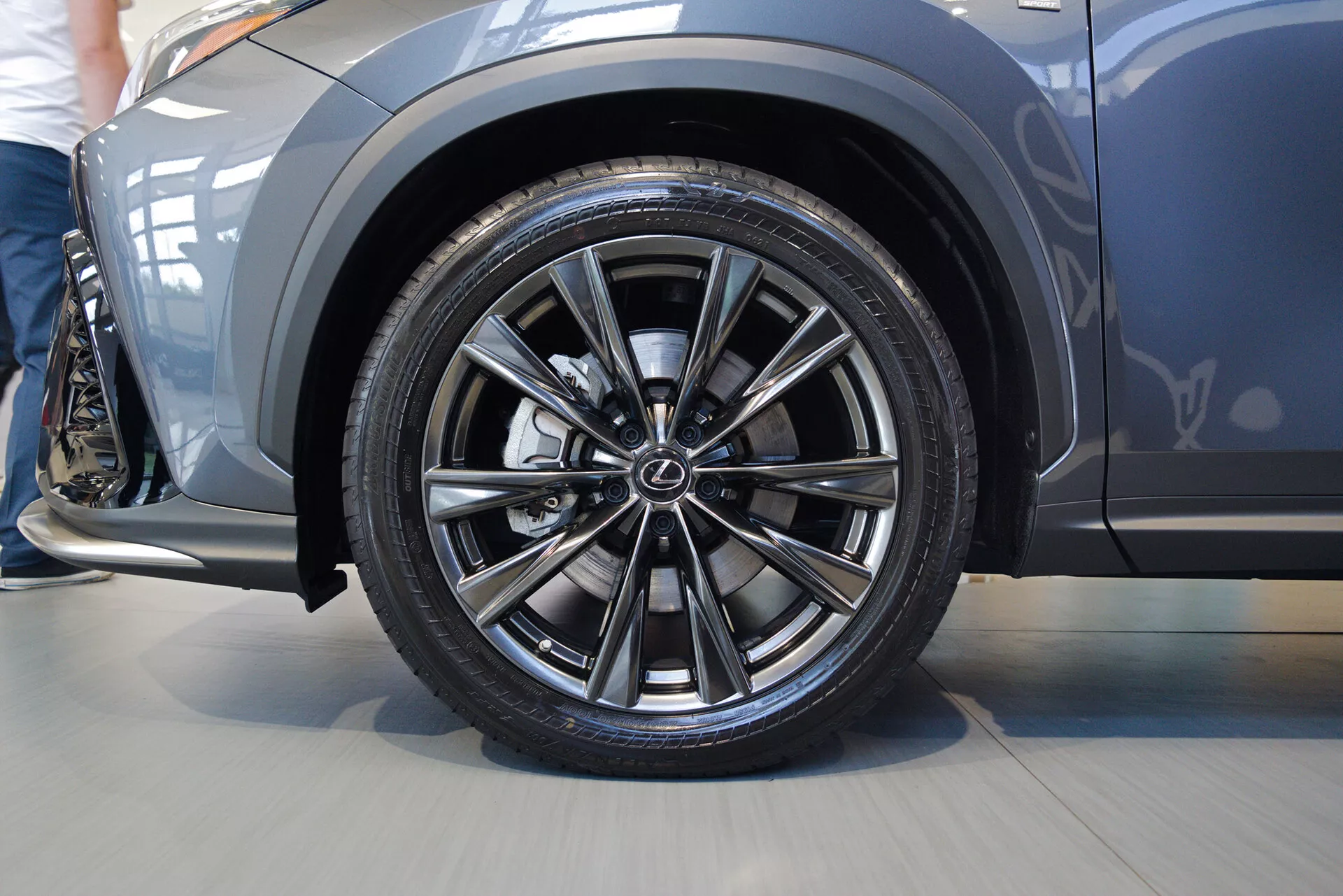 Gros plan en contre-plongée de la roue avant côté conducteur d'une Lexus NX 2021, mettant en avant la jante en alliage avec un design à rayons jumelés et le logo du fabricant. Le pneu noir à profil bas met en évidence les performances sportives. L'aile au-dessus de la roue est métallique et légèrement bombée, indiquant un look sportif. Le panneau latéral montre un design aérodynamique élégant. Image prise en intérieur avec une grande proximité, centrée sur la roue et le panneau latéral avant.