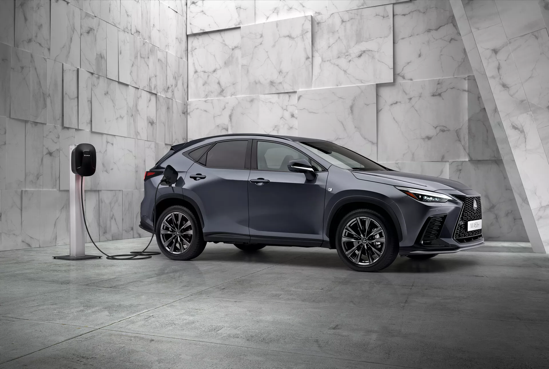 Cette image présente le Lexus NX 2021, un SUV compact moderne avec un design élégant et sportif, photographié sous un angle trois-quarts avant. Le côté gauche du véhicule et partiellement son avant sont visibles, positionnés en diagonale dans un cadre minimaliste orné de grandes tuiles de marbre clair, suggérant une ambiance premium. Le Lexus NX affiche des phares LED angulaires distinctifs et une calandre avant proéminente, mettant en avant sa nature sophistiquée. On observe des rétroviseurs latéraux aérodynamiques, des jantes en alliage à rayons multiples complexes et des poignées de porte encastrées qui soulignent le design sportif et avancé. Un détail crucial est la connexion de la voiture à une station de recharge électrique, indiquant qu'il s'agit d'un véhicule électrique ou hybride rechargeable. L'aile avant comprend une prise de recharge, indiquant les capacités électriques du véhicule. Cette image statique se concentre sur la présentation du design du véhicule et de ses caractéristiques technologiques dans une composition équilibrée et calme avec un ample espace négatif, symbolisant l'exclusivité et l'innovation dans le design automobile.