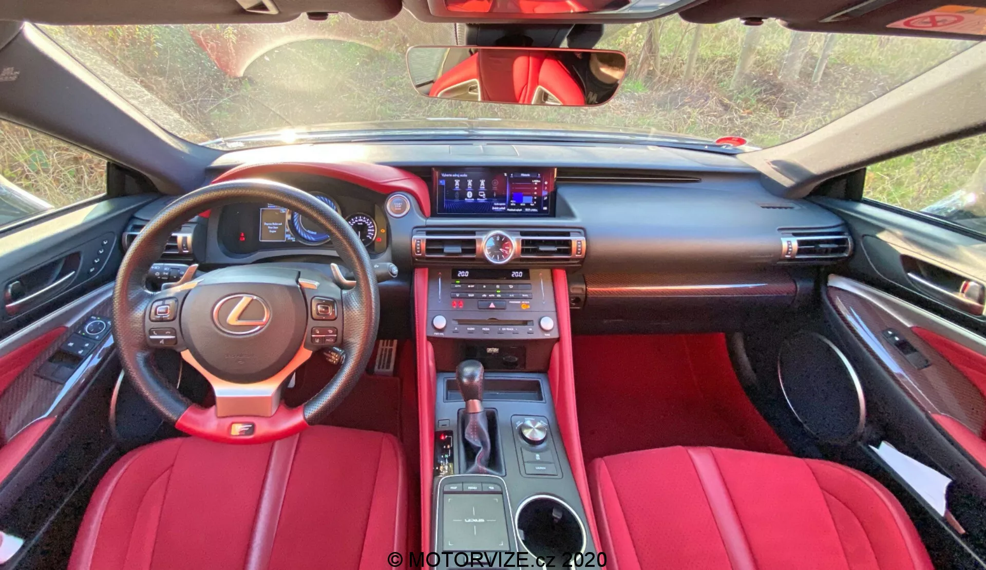 Vue intérieure de la Lexus RC 2016 depuis une perspective en première personne. L'image est prise depuis l'espace des sièges arrière vers l'avant, capturant l'ensemble de la cabine avant. Une garniture rouge riche et noire est visible au premier plan en haut, probablement des sièges arrière ou une cloison intérieure. Le tableau de bord expansif, orné de matériaux de haute qualité en noir et rouge, présente des coutures visibles, des bouches d'aération centrales avec des accents argentés et un écran d'infodivertissement affichant des médias ou de la navigation. En dessous se trouvent les boutons de contrôle du climat, d'autres commandes sur la console centrale, le système audio et possiblement des fonctionnalités de chauffage/refroidissement des sièges. Le volant gainé de cuir avec l'emblème Lexus montre des commandes multifonctions. Le tableau de bord est partiellement visible. La console centrale comprend un levier de vitesse automatique, un pavé tactile ou une molette et des porte-gobelets. L'intérieur exsude le luxe avec des fonctionnalités multimédias modernes et une technologie avancée.