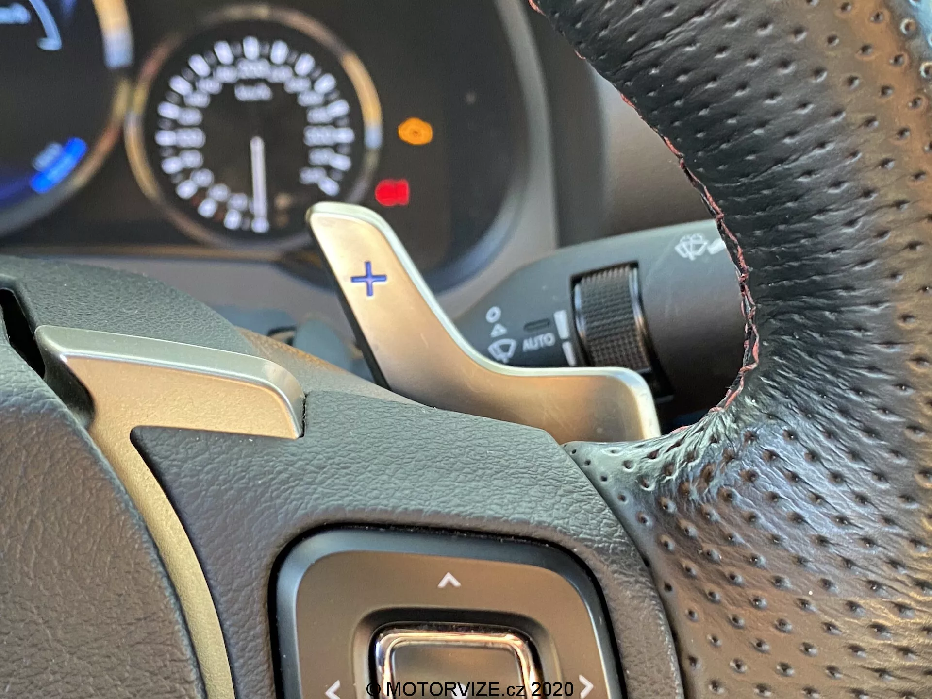 Vue en gros plan, perspective du conducteur, du volant de la Lexus RC 2016 avec prise texturée, possiblement en cuir, accent chromé, bouton d'assistance à la conduite sur le côté gauche, palette de changement de vitesse métallique derrière le côté droit du volant avec signe '+' pour des montées de rapports manuelles, et tableau de bord partiellement visible suggérant un moteur sportif avec un compteur de tours atteignant les 8000 RPM ; mise au point intérieure avec arrière-plan flou mettant en évidence les détails du volant et de la palette de changement.