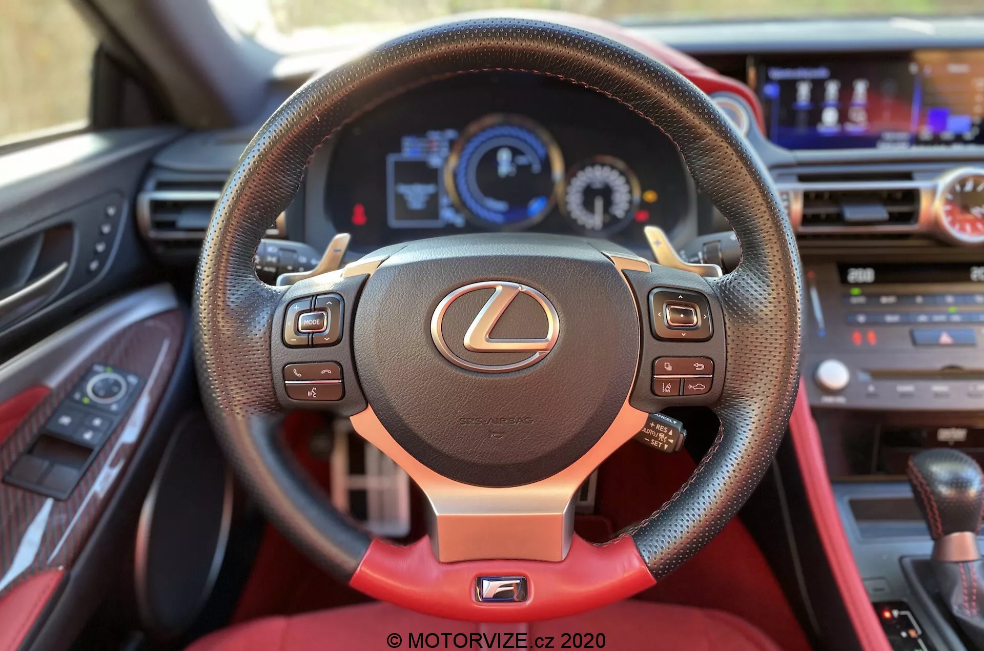 Vue intérieure depuis le siège du conducteur d'une Lexus RC 2016, mettant en avant le volant avec l'emblème Lexus. Le volant à trois branches comprend des commandes multifonctions, du cuir avec des surpiqûres rouges et un accent rouge sur la branche inférieure. Le tableau de bord numérique derrière le volant affiche un compteur de vitesse, un compte-tours et un écran multifonction. La partie supérieure du tableau de bord inclut un système d'infodivertissement et des boutons de commande de climatisation. Une partie de la console centrale avec le sélecteur de vitesse est visible sous le tableau de bord.