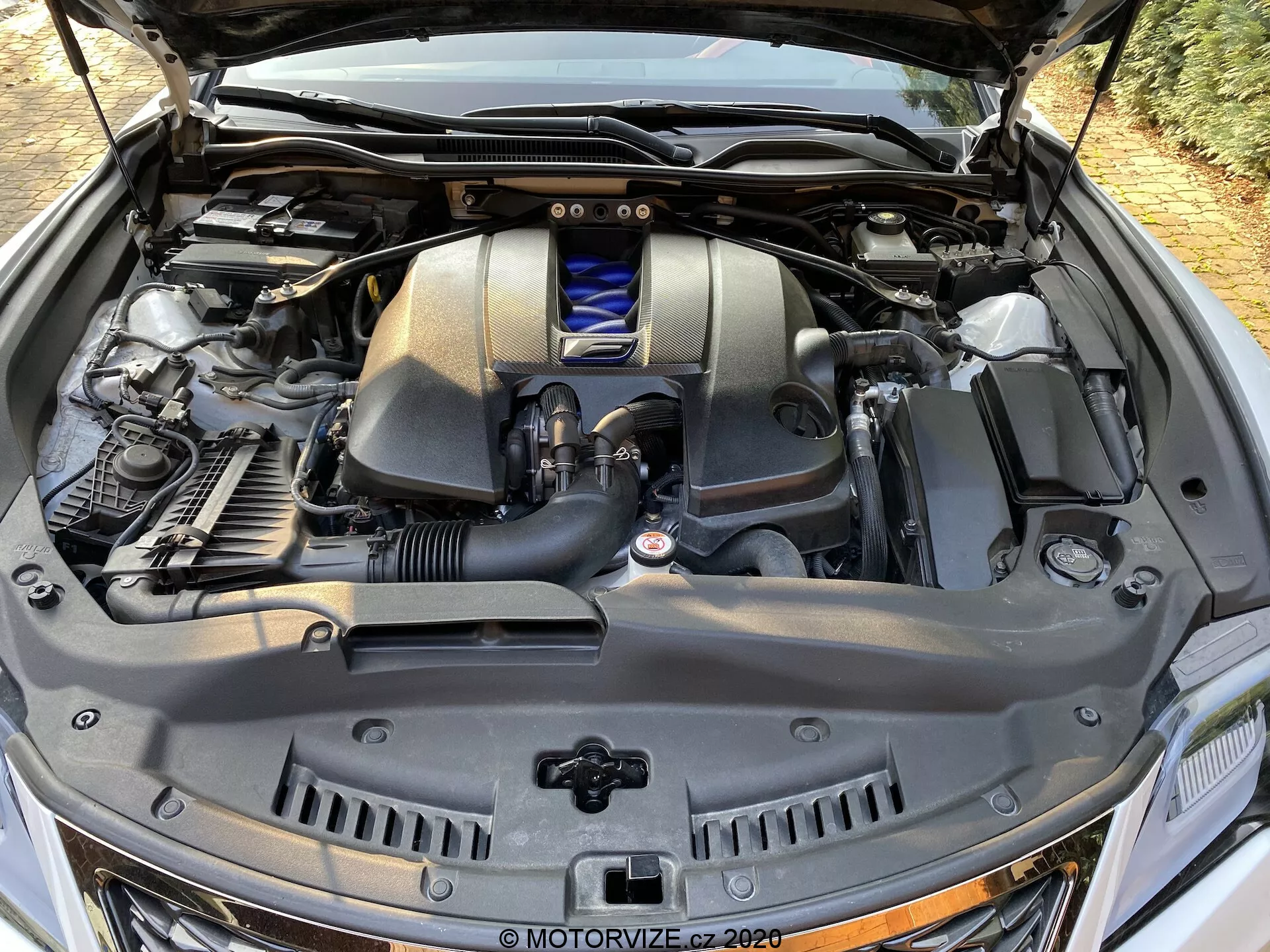Vue rapprochée et détaillée du compartiment moteur de la Lexus RC 2016 depuis un angle avant-droit, mettant en évidence le cache-moteur avec emblème, le système d'admission d'air, les réservoirs de liquide avec bouchon bleu, les tuyaux métalliques et en caoutchouc, le dessus du radiateur et les composants structurels se détachant sur le fond de la cloison pare-feu et des modules électroniques de la voiture.