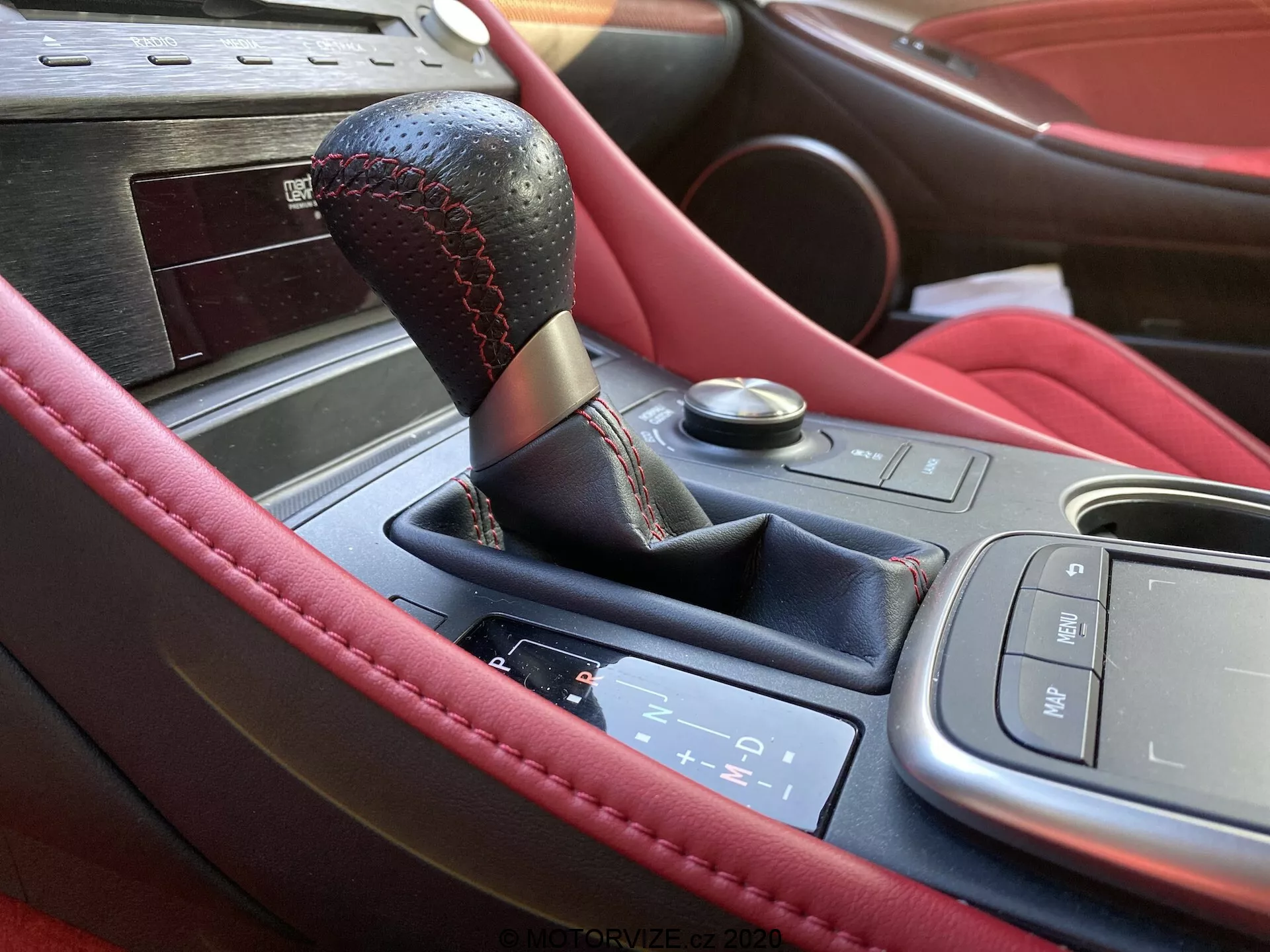 Vue rapprochée de la console centrale de l'intérieur de la Lexus RC 2016, mettant en avant un levier de vitesses en cuir noir avec des coutures rouges, un panneau de changement de vitesse électronique pour une transmission automatique, une interface de contrôle multimédia, un bouton de démarrage/arrêt du moteur et des garnitures en bois luxueuses. Les caractéristiques incluent des matériaux en cuir de qualité supérieure et probablement des porte-gobelets, suggérant un design sportif et luxueux.