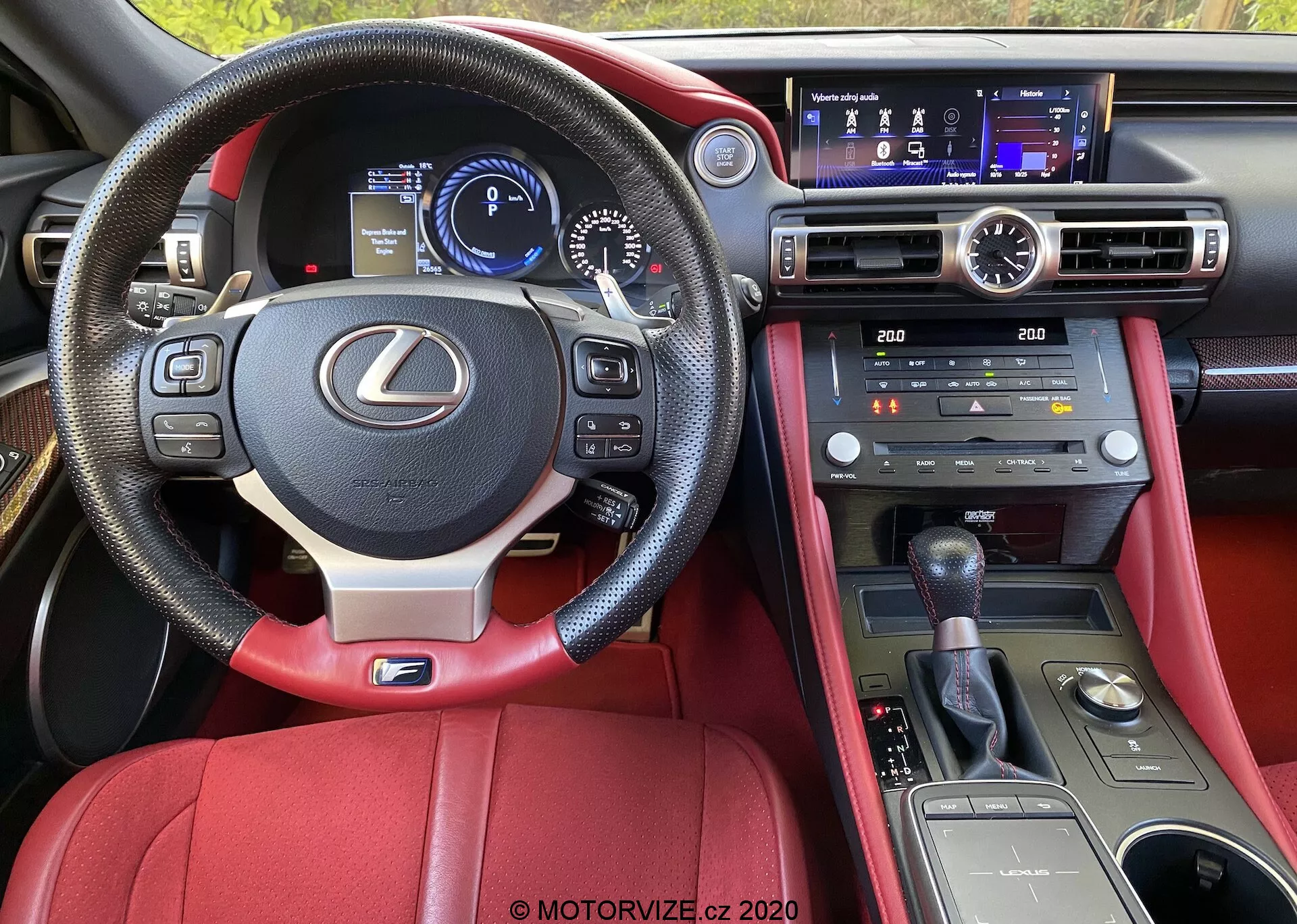 Vue intérieure d'une Lexus RC 2016 depuis la perspective du conducteur, mettant en avant le volant en cuir avec commandes intégrées, le tableau de bord numérique, la console centrale avec écran multifonction et commandes de climatisation, le levier de vitesse manuel, la sellerie en cuir rouge et les caractéristiques technologiques avancées.