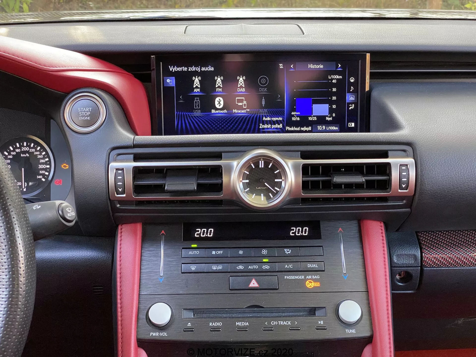 L'image capture l'intérieur d'une Lexus RC 2016 depuis la perspective de l'un des sièges avant, mettant en vedette un écran tactile multifonction moderne au centre du tableau de bord. Au-dessus de l'écran se trouve une élégante horloge analogique. Le tableau de bord comprend des bouches d'aération verticales de chaque côté de l'écran, des lattes horizontales réglables pour le flux d'air, et de nombreux contrôles physiques et boutons pour le contrôle de la climatisation, le chauffage des sièges, les fonctions média et radio. Le petit affichage de la climatisation indique un système de contrôle de la climatisation à deux zones. Le mélange de détails en cuir rouge et de matériau texturé noir ajoute à l'esthétique sportive et de haute qualité, tandis que l'éclairage d'ambiance apporte une touche raffinée.